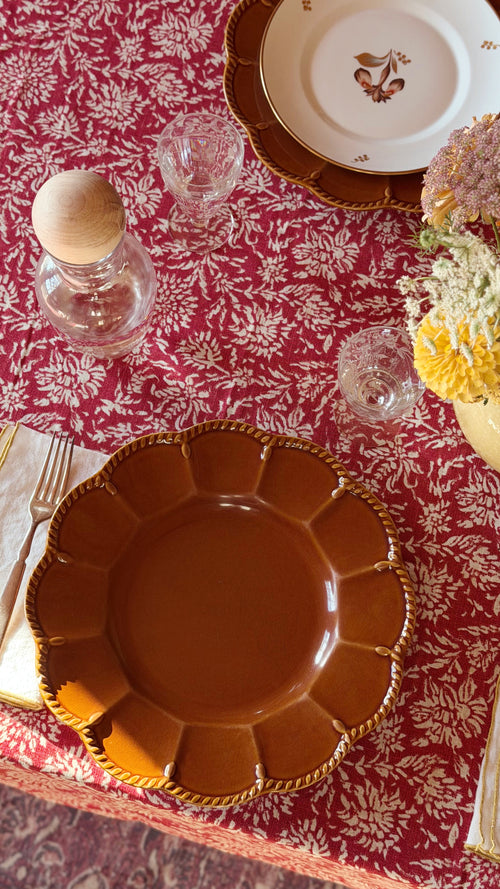 An overhead view of a styled table setting featuring aVintage Certified International Florentine Brown Dinner Plate by Kari Walmsley with a scalloped rim and gold rope detailing, placed on a red and white floral tablecloth alongside glassware, a small bottle, and floral accents.