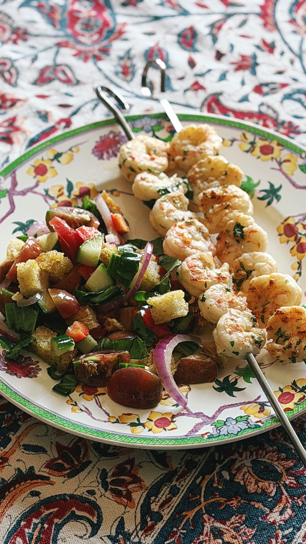 Shrimp skewers and a colorful vegetable salad on a decorative floral plate, set on a patterned tablecloth.