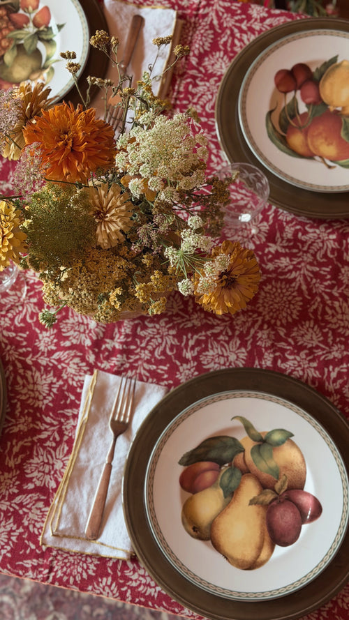 An overhead view of a styled dining table setting featuring a red and white patterned tablecloth, ceramic plates with hand-painted fruit illustrations, a folded cream napkin with a gold fork, and a central dried flower arrangement.