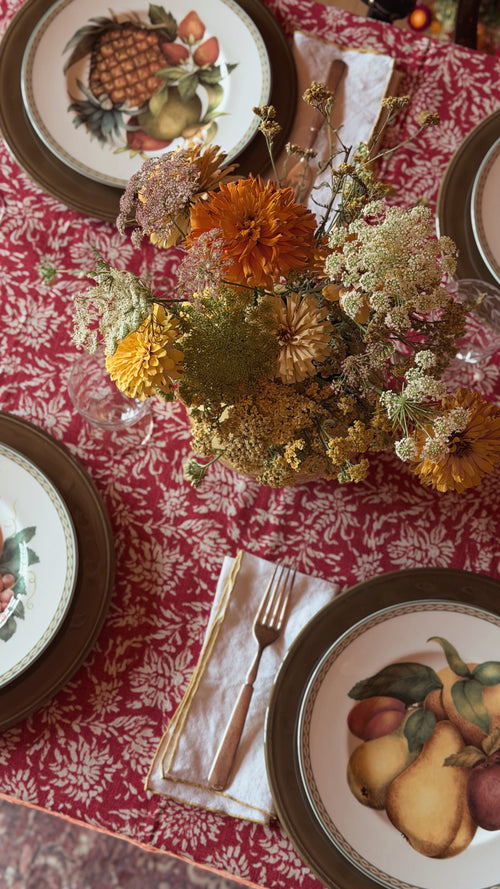 An overhead view of a styled dining table setting featuring a red and white damask tablecloth, a central dried flower arrangement, Vintage Fitz & Floyd Belle Classique Fruit Salad Plates, gold-toned cutlery, and folded napkins.