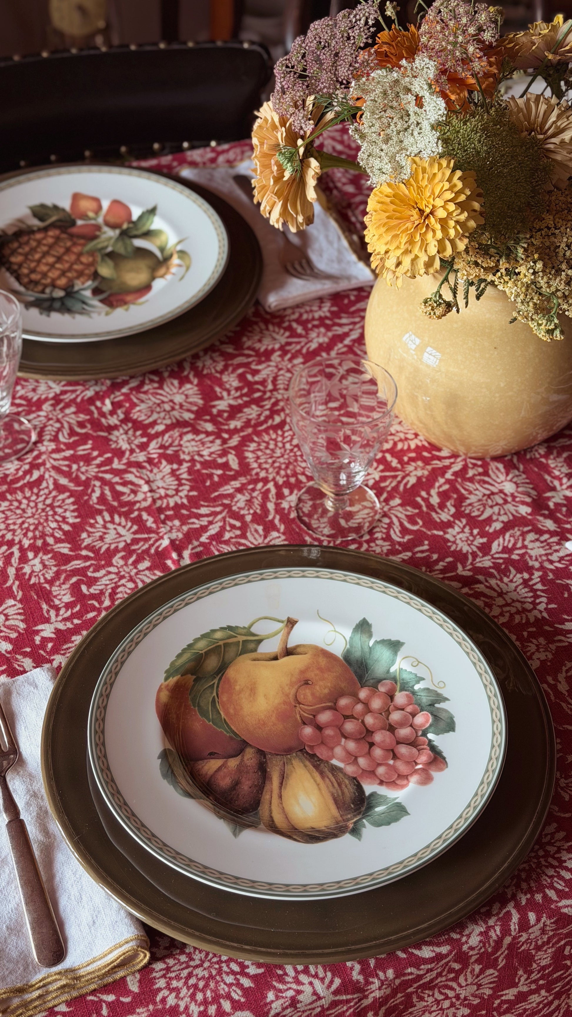 A styled dining table setting featuring a white ceramic plate with a colorful fruit illustration (pears, grapes, plums) and a dark brown charger plate, set on a red and white damask tablecloth next to a floral vase.