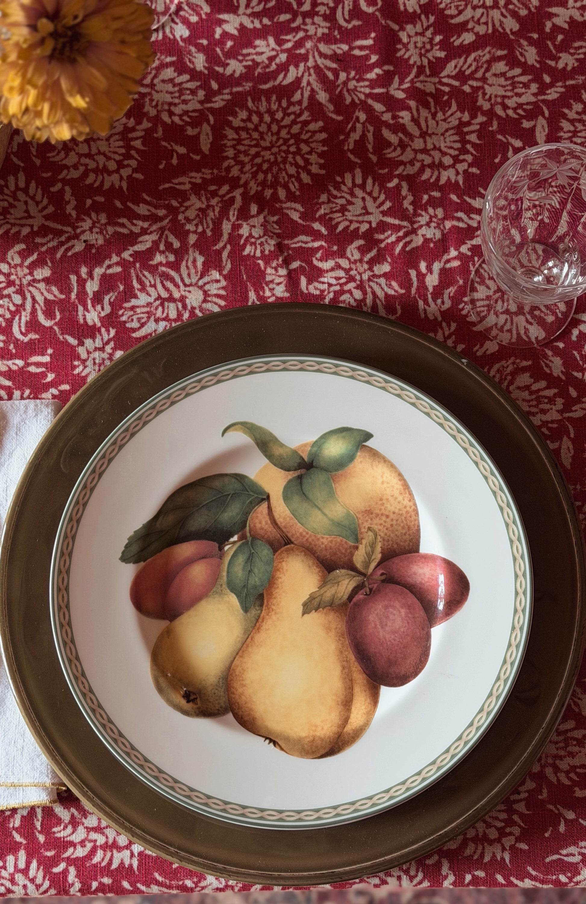 A Vintage Fitz & Floyd Belle Classique Fruit Salad Plate with a painted fruit motif, placed on a dark brown charger plate over a red damask tablecloth. A glass and a yellow flower are partially visible in the frame.