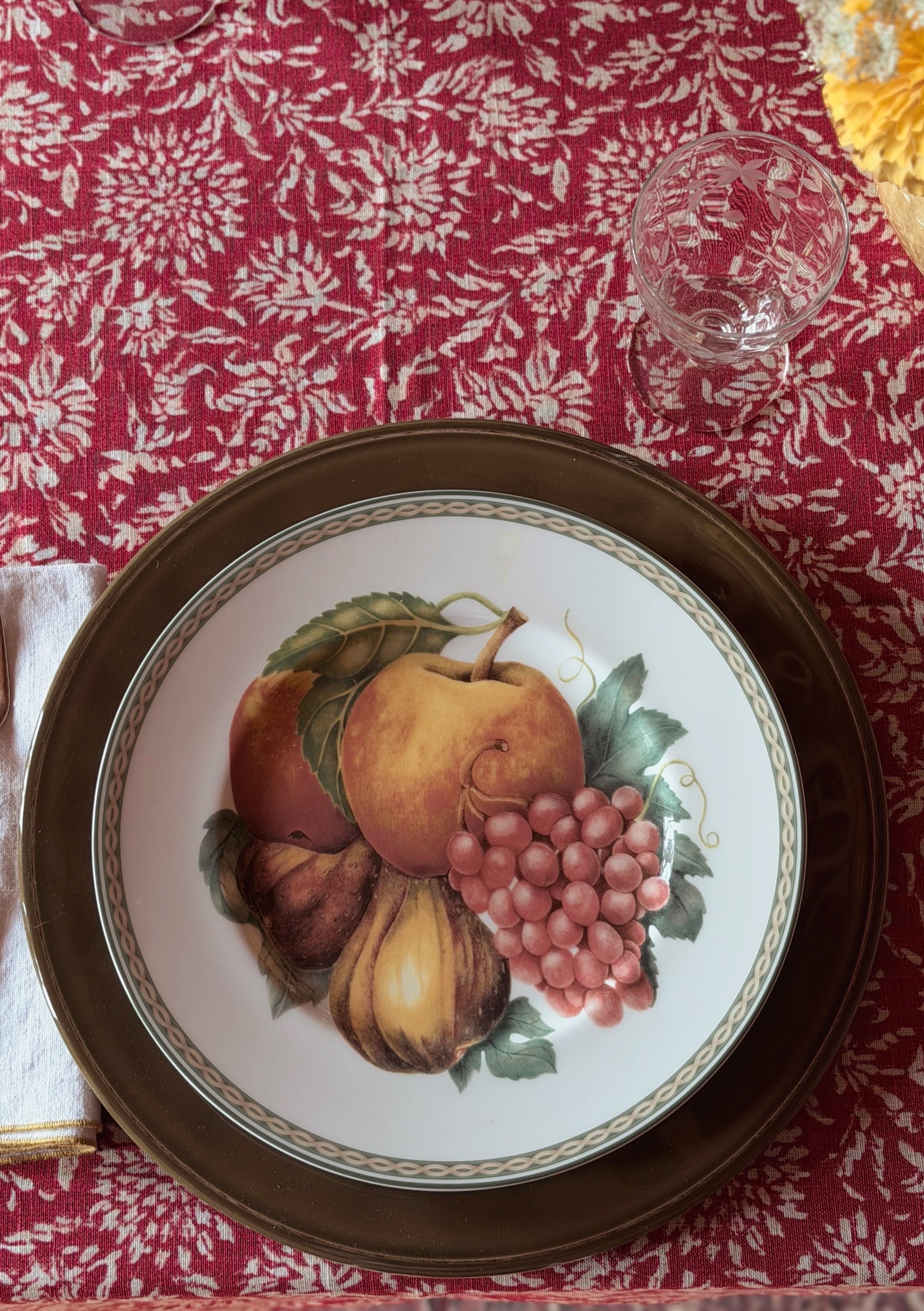 A Vintage Fitz & Floyd Belle Classique Fruit Salad Plate with a painted fruit motif, placed on a matching dark brown charger plate over a red and white patterned tablecloth. A clear glass is partially visible at the top right.