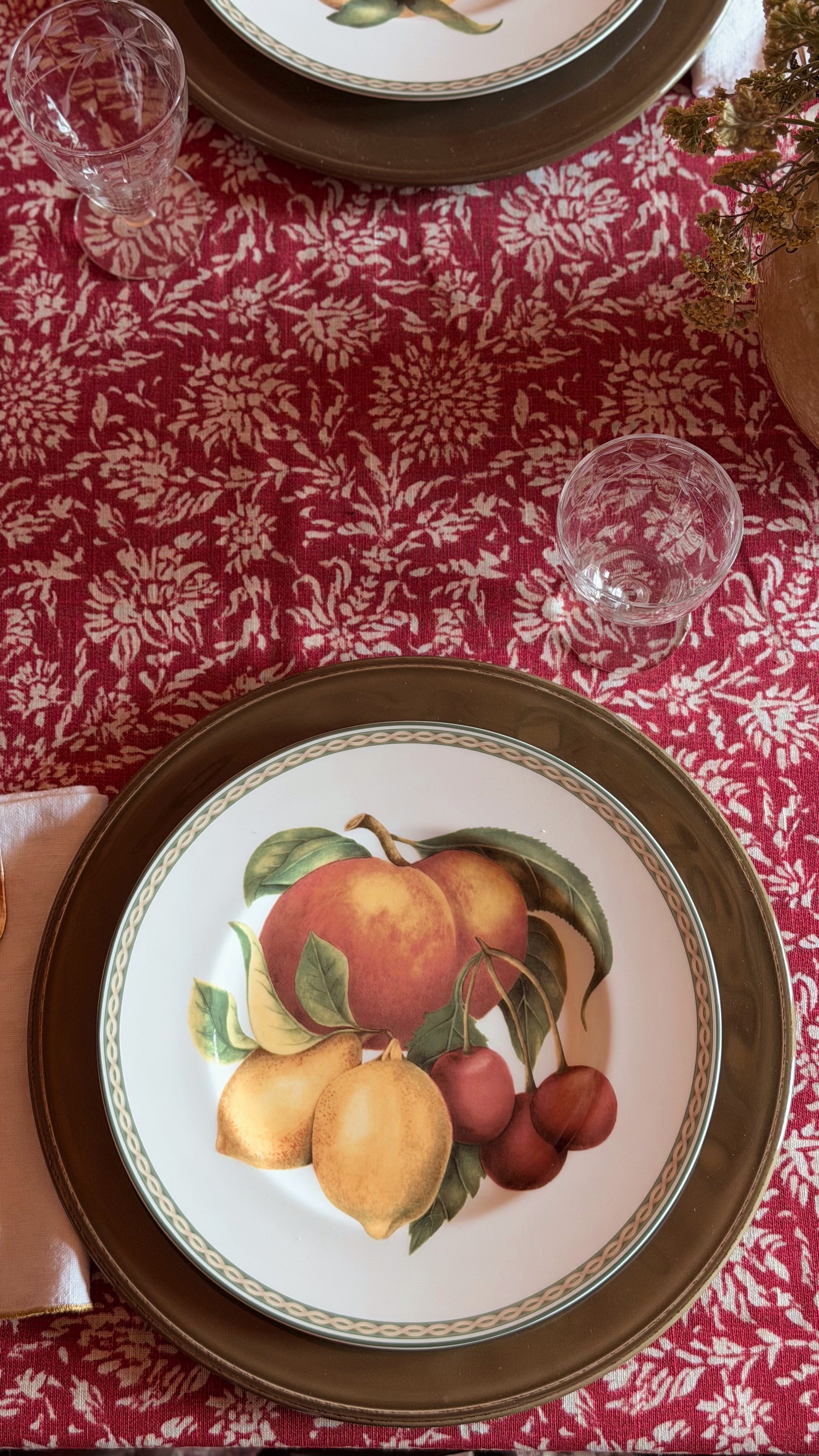 A close-up overhead view of a place setting featuring a Vintage Fitz & Floyd Belle Classique Fruit Salad Plate with a painted fruit illustration of peaches, plums, and green leaves, surrounded by a brown charger plate and a red-and-white patterned tablecloth.