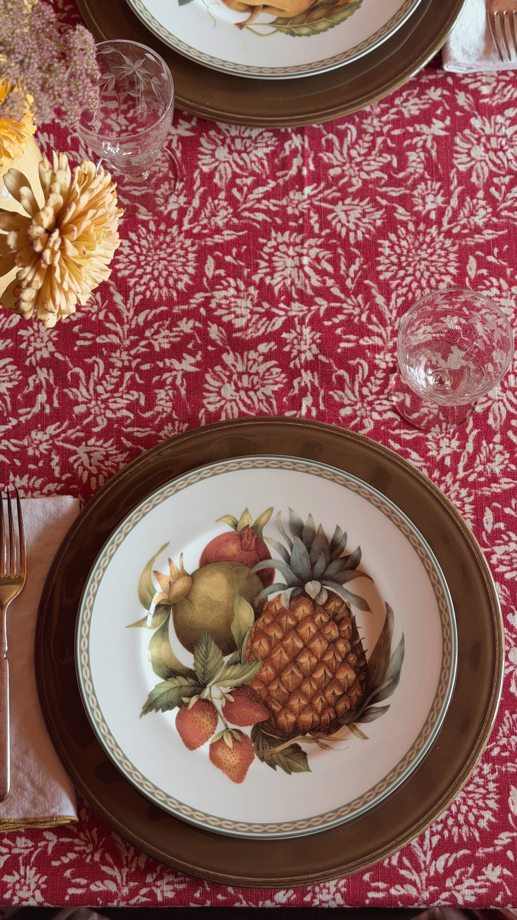 A styled tabletop setting featuring a red and white damask tablecloth, a Vintage Fitz & Floyd Belle Classique Fruit Salad Plate with a fruit illustration (pineapple, pear, strawberries), and gold-rimmed charger plates.
