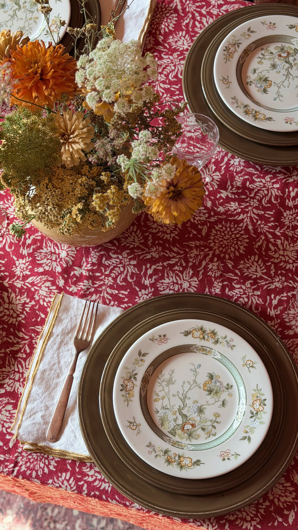 A styled tabletop setting with a red and white patterned tablecloth, stacked plates with a floral center plate, a folded napkin with a fork, and a dried flower arrangement in a vase.