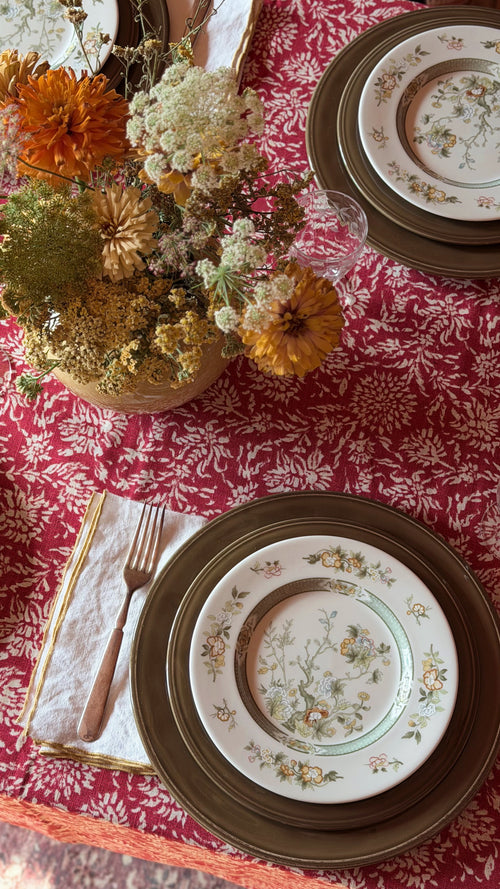 A styled tabletop setting with a red and white patterned tablecloth, stacked plates with a floral center plate, a folded napkin with a fork, and a dried flower arrangement in a vase.
