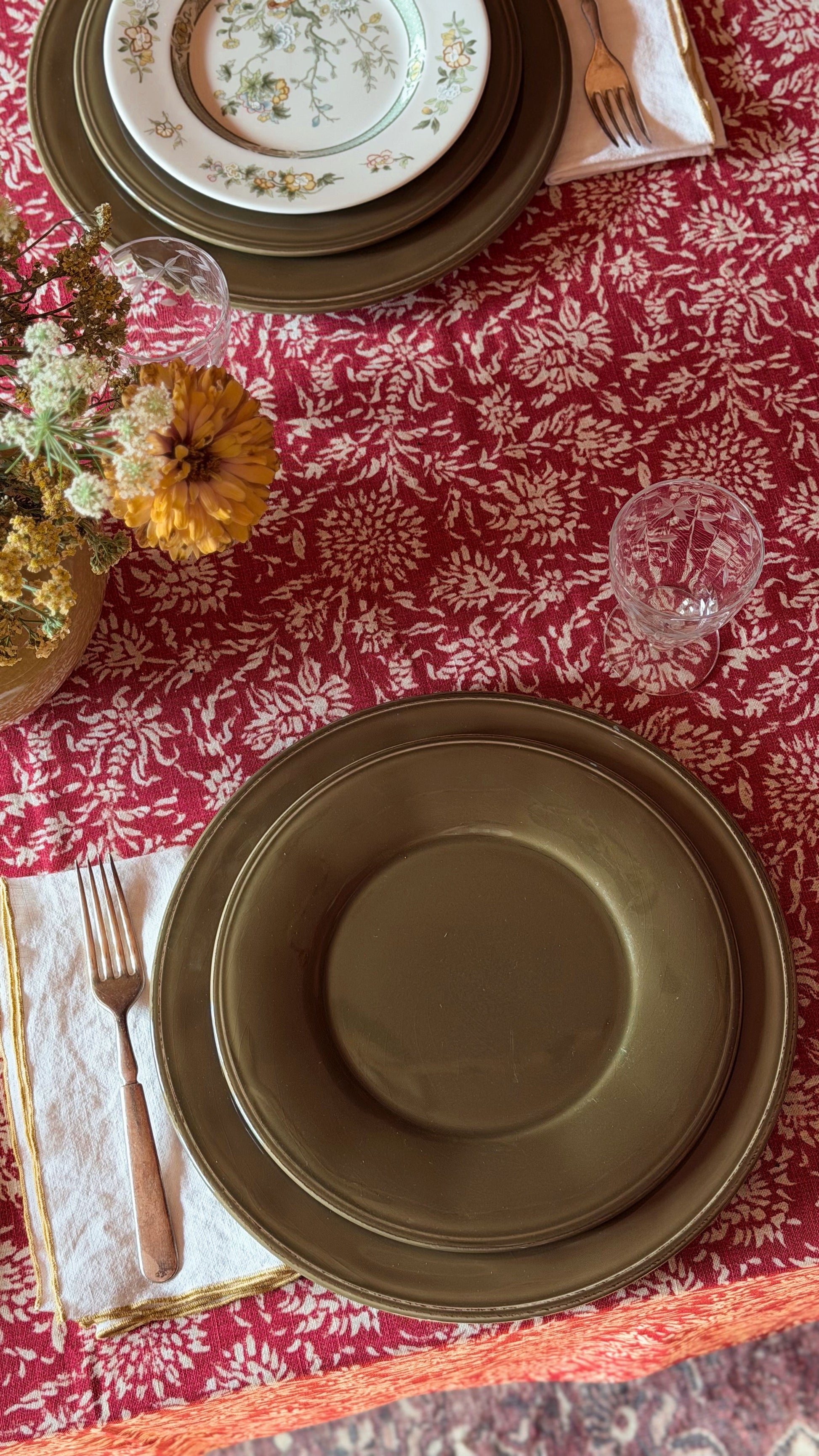 An overhead view of a styled table setting featuring dark olive green ceramic plates, a red and white floral tablecloth, gold-toned cutlery on cream napkins, and a small floral arrangement.