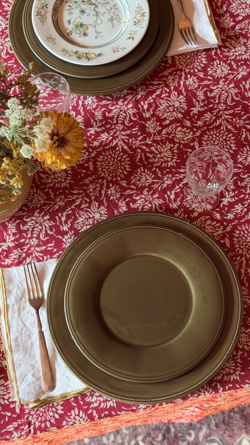 An overhead view of a styled table setting featuring dark olive green ceramic plates, a red and white floral tablecloth, gold-toned cutlery on cream napkins, and a small floral arrangement.