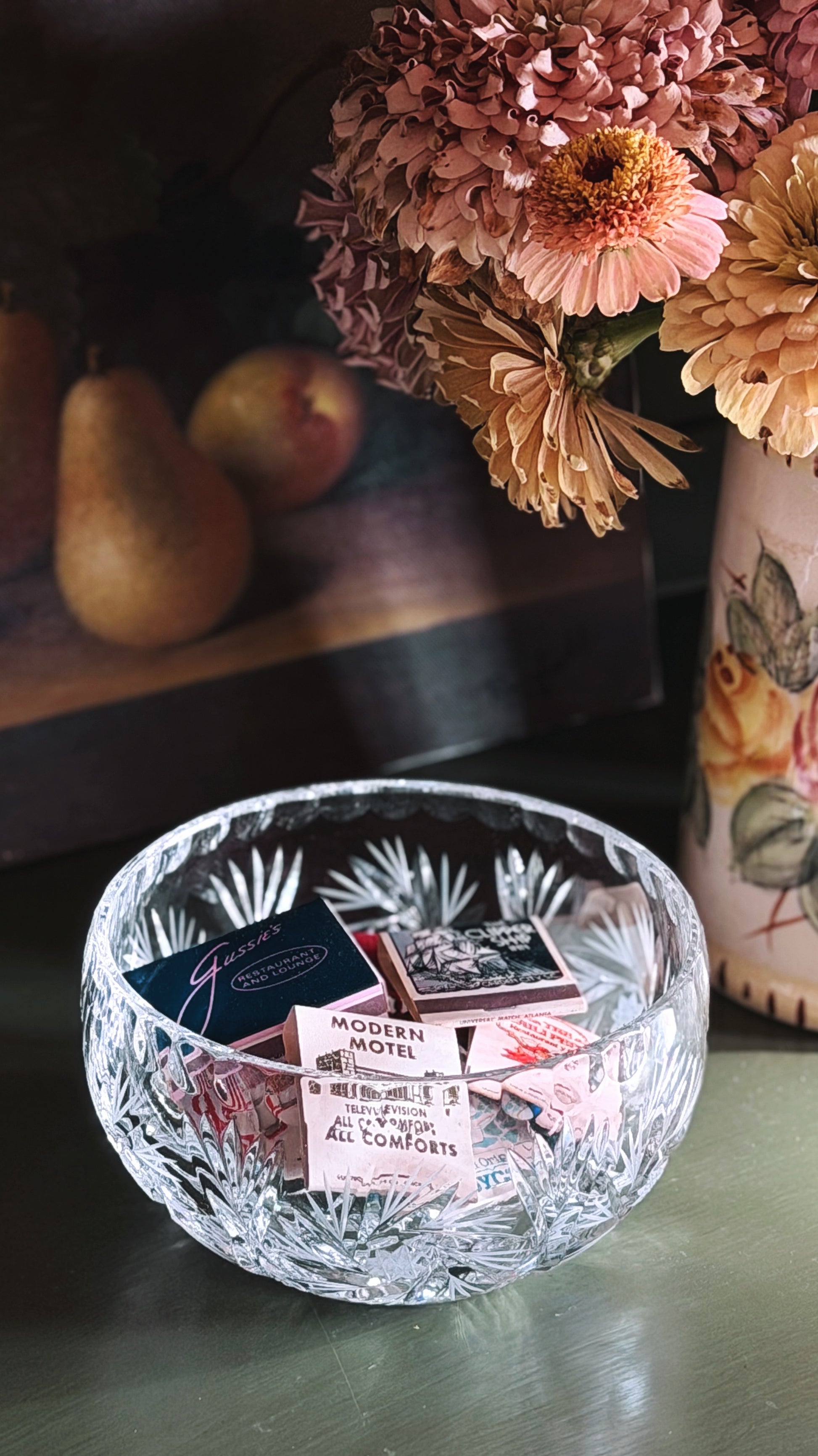 A clear cut-crystal bowl with a faceted starburst pattern sits on a dark tabletop, holding small vintage matchbooks. In the softly blurred background, dried pink flowers in a vase and pears are visible.