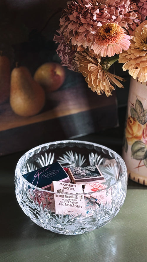 A clear cut-crystal bowl with a faceted starburst pattern sits on a dark tabletop, holding small vintage matchbooks. In the softly blurred background, dried pink flowers in a vase and pears are visible.
