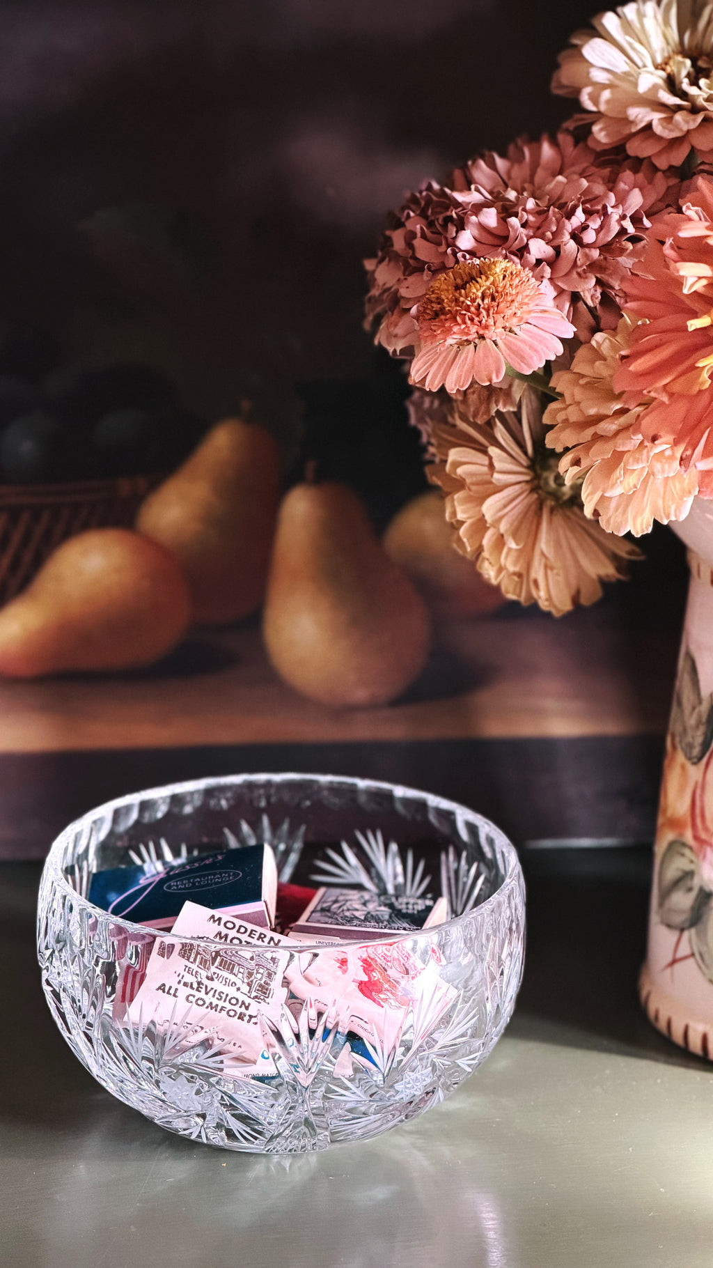 A clear cut-crystal bowl filled with colorful matchbooks, set on a tabletop in front of a floral still life background with pears and pink flowers.