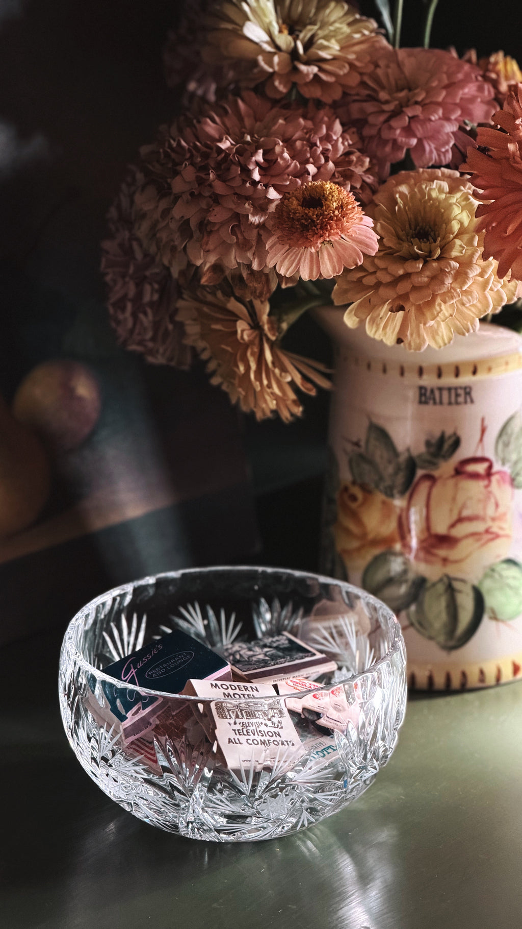 A clear cut-crystal bowl filled with small matchbooks, placed on a tabletop in a styled interior scene with dried flowers and a floral canister in the background.