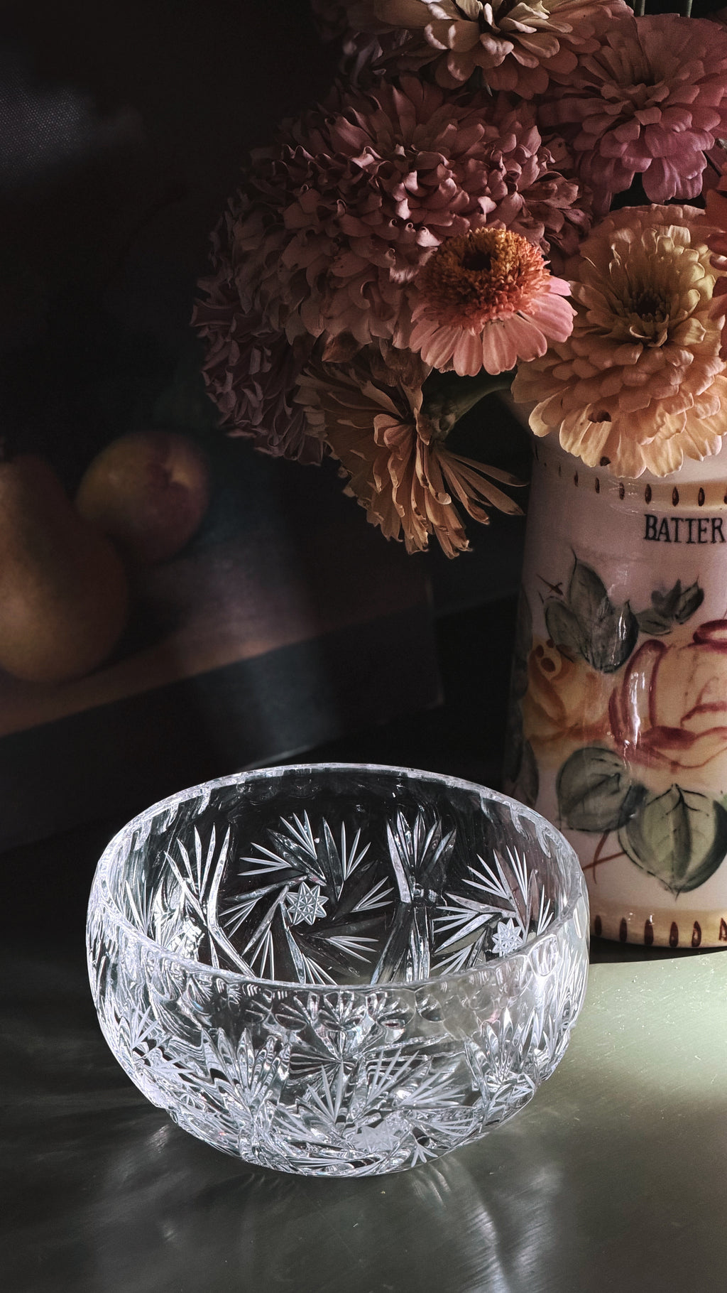 A clear cut-crystal bowl with a floral pattern, set on a reflective surface with a dark background and dried flowers in a vase behind it.