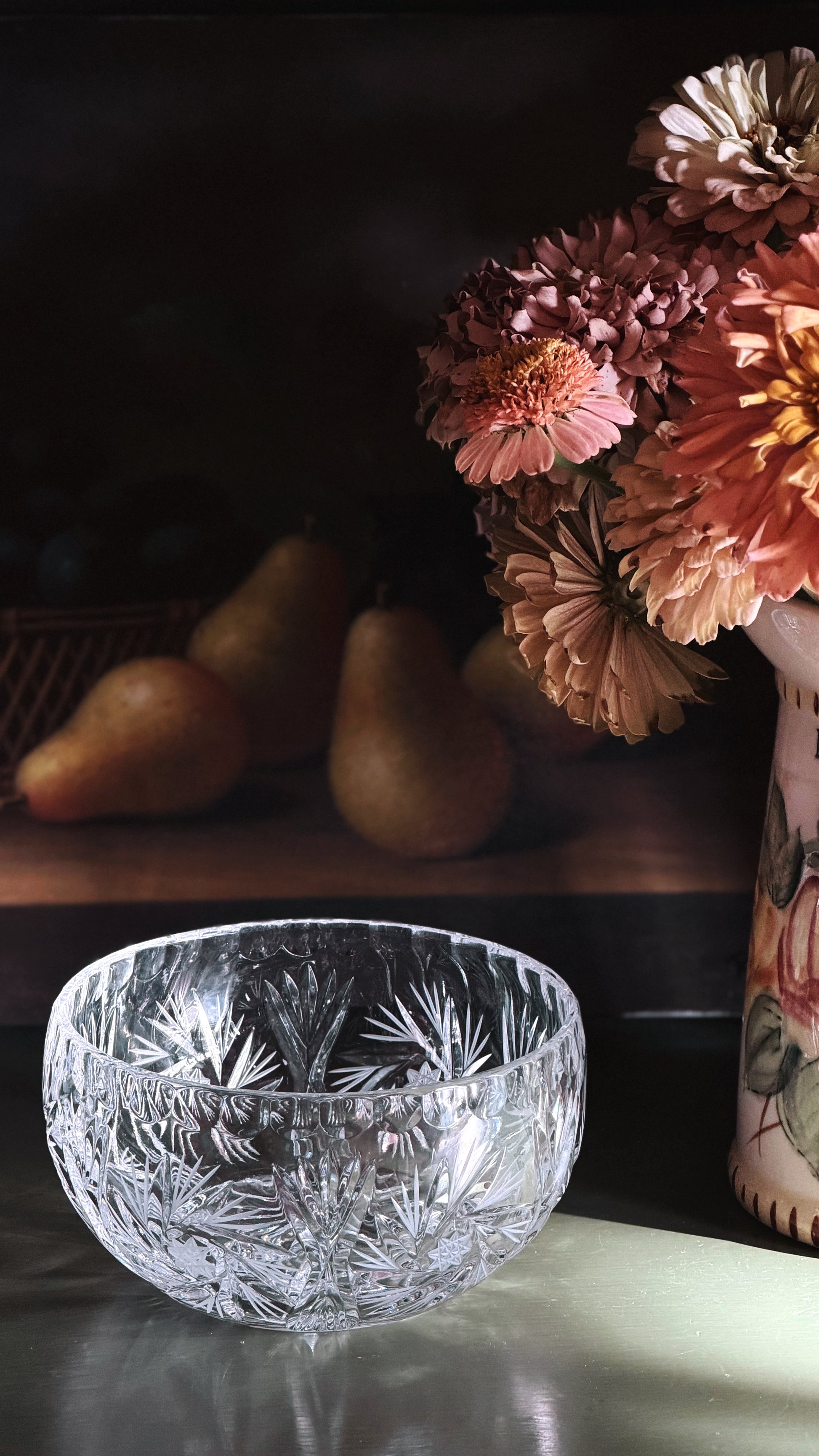 A clear cut-crystal bowl with a floral pattern sits on a reflective surface in a dimly lit setting, with pears blurred in the background and dried flowers in a vase to the right.
