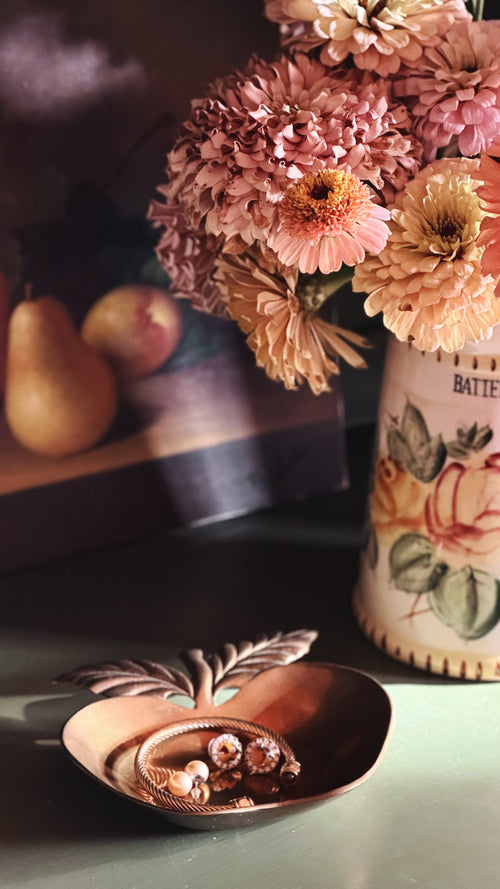 A styled tabletop scene with a copper-toned decorative bowl in the foreground holding small jewelry pieces, next to a ceramic vase filled with pink and peach dried flowers. A dark box and two pears are blurred in the background.