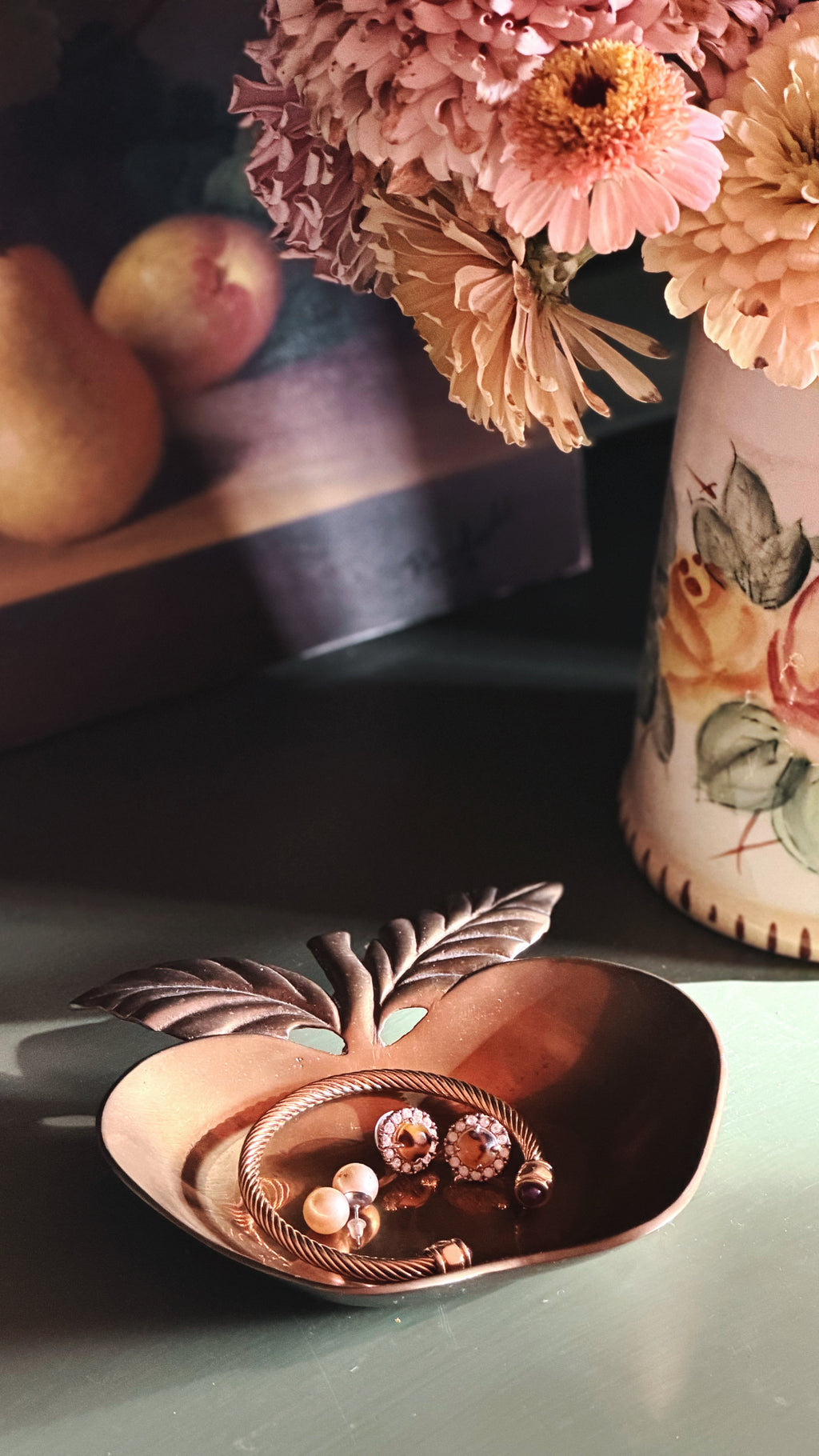 A brass apple-shaped jewelry tray holding a gold ring and earrings, styled on a tabletop with dried flowers and fruit in the background.
