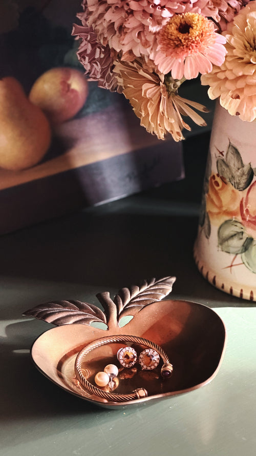 A brass apple-shaped jewelry tray holding a gold ring and earrings, styled on a tabletop with dried flowers and fruit in the background.