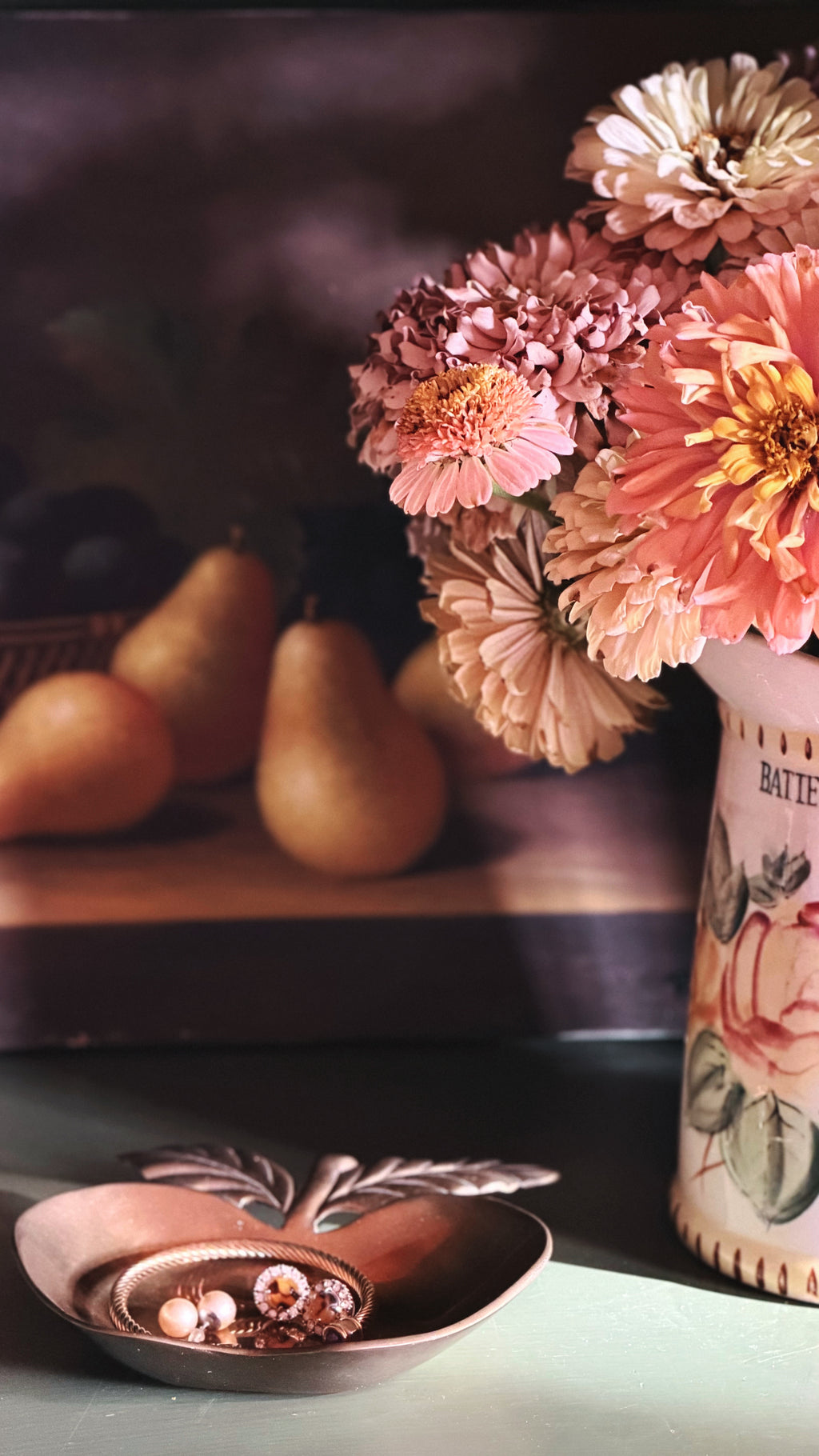 A styled still life scene with a ceramic vase holding pink, peach, and cream flowers, next to a small brass dish with decorative leaves. In the softly blurred background, pears rest on a surface against a dark backdrop.