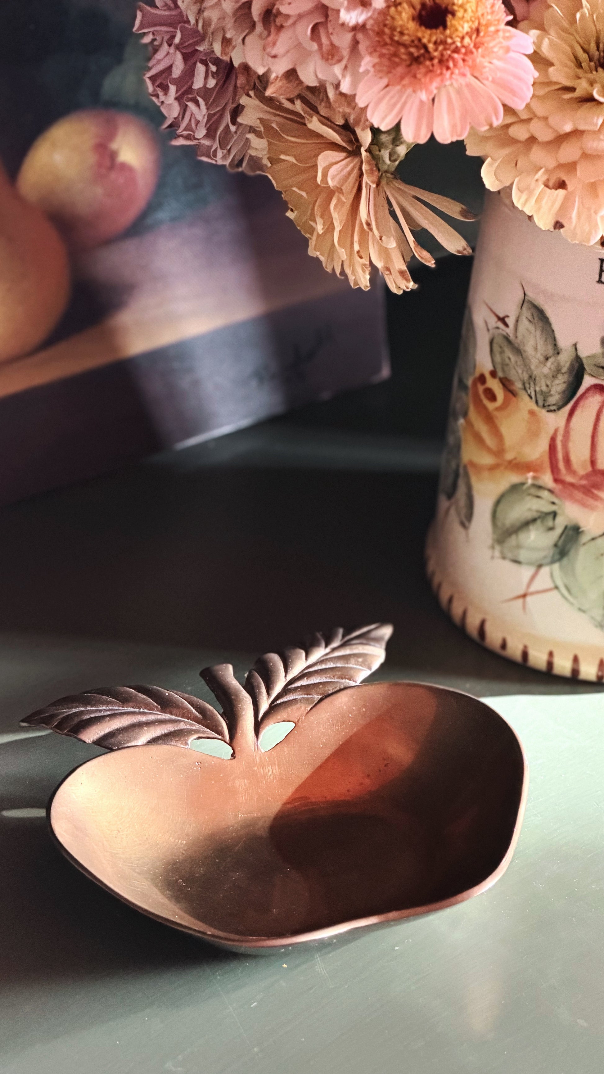 An apple-shaped brass dish with leaf handles, styled on a tabletop next to a floral vase and blurred fruit.