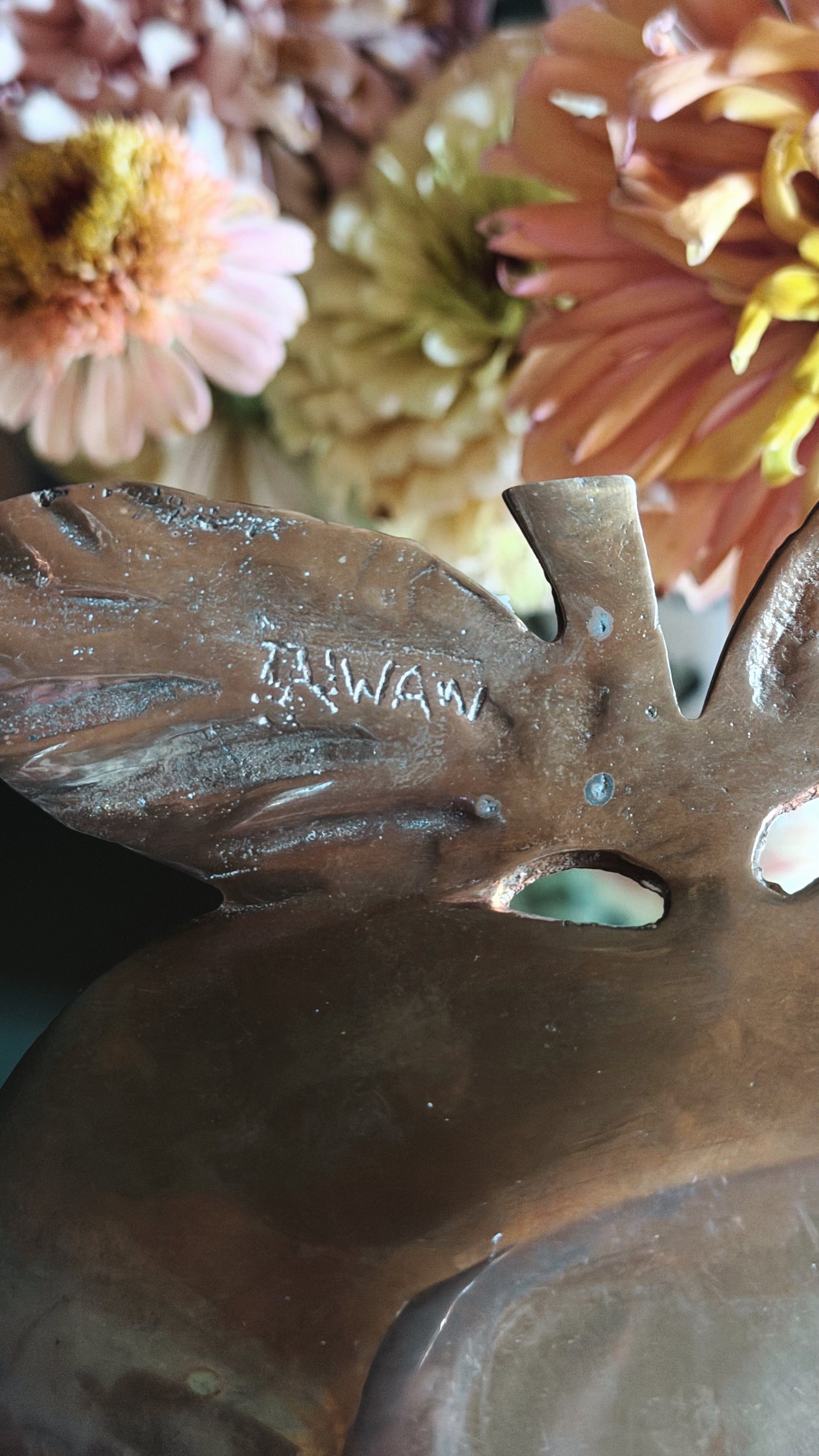 Close-up of a bronze-toned decorative object with a leaf-like shape and the word 'TAIWAN' engraved on it, set against a blurred background of dried flowers.
