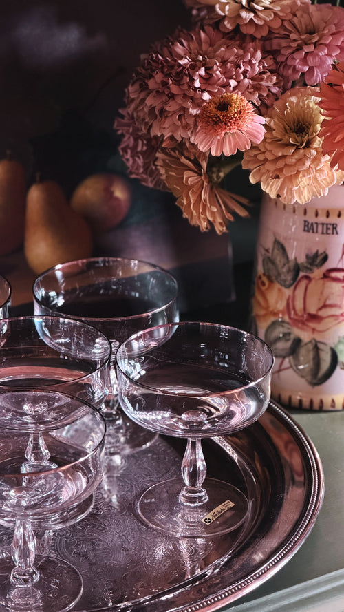 A set of clear glass coupe-style drinking glasses arranged on a round silver tray, with a floral arrangement and fruit in the background.