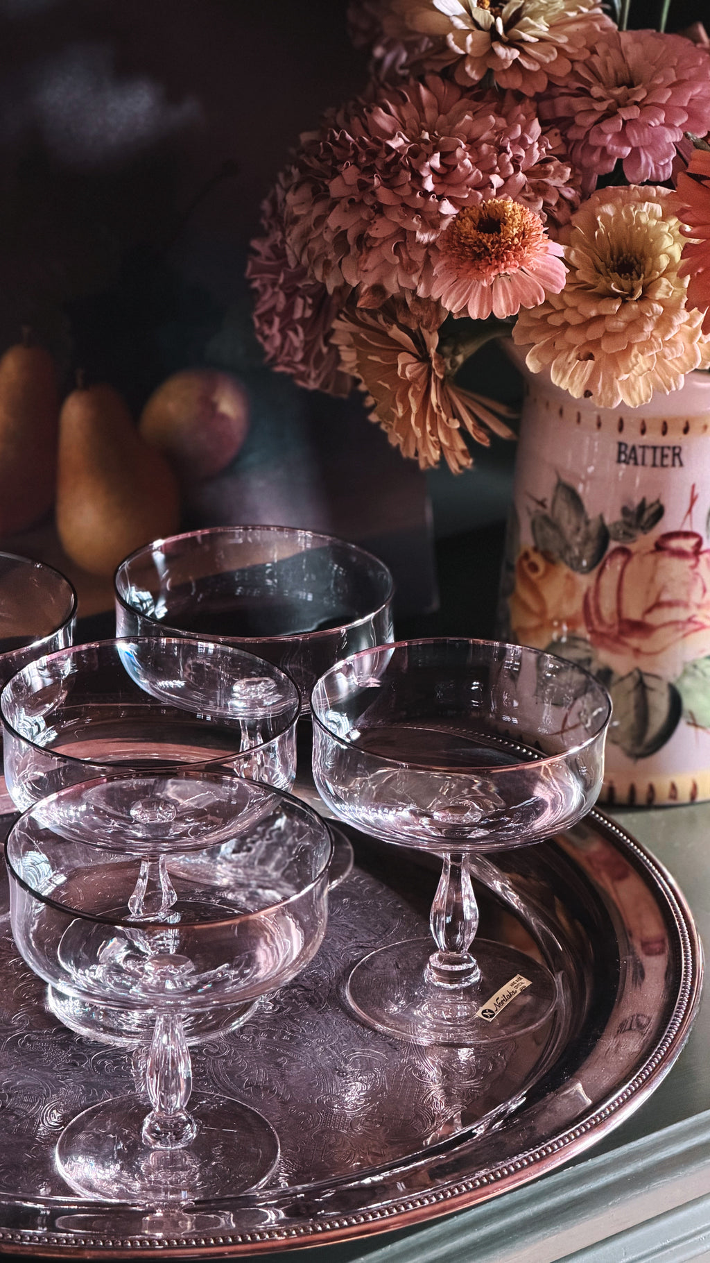 A set of crystal coupe glasses arranged on a round silver tray, with a blurred floral arrangement and pears in the background.