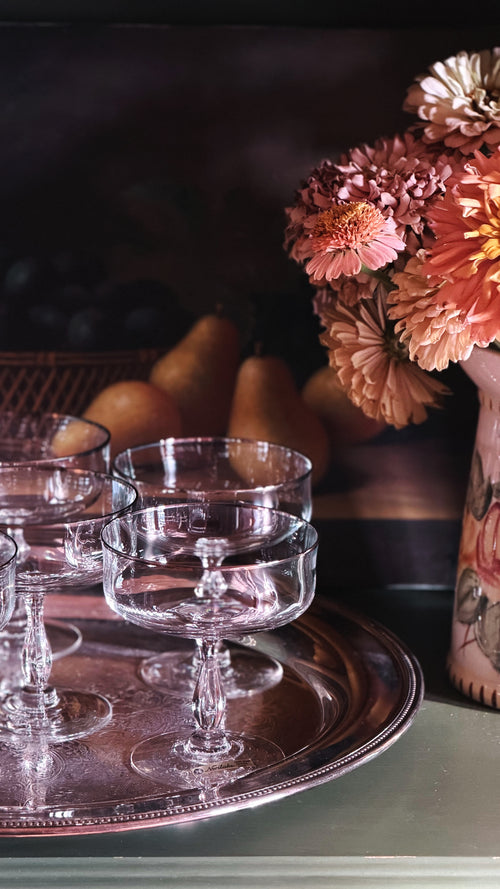 Crystal coupe-style glasses stacked on a round silver tray, with a blurred floral arrangement and pears in the background.