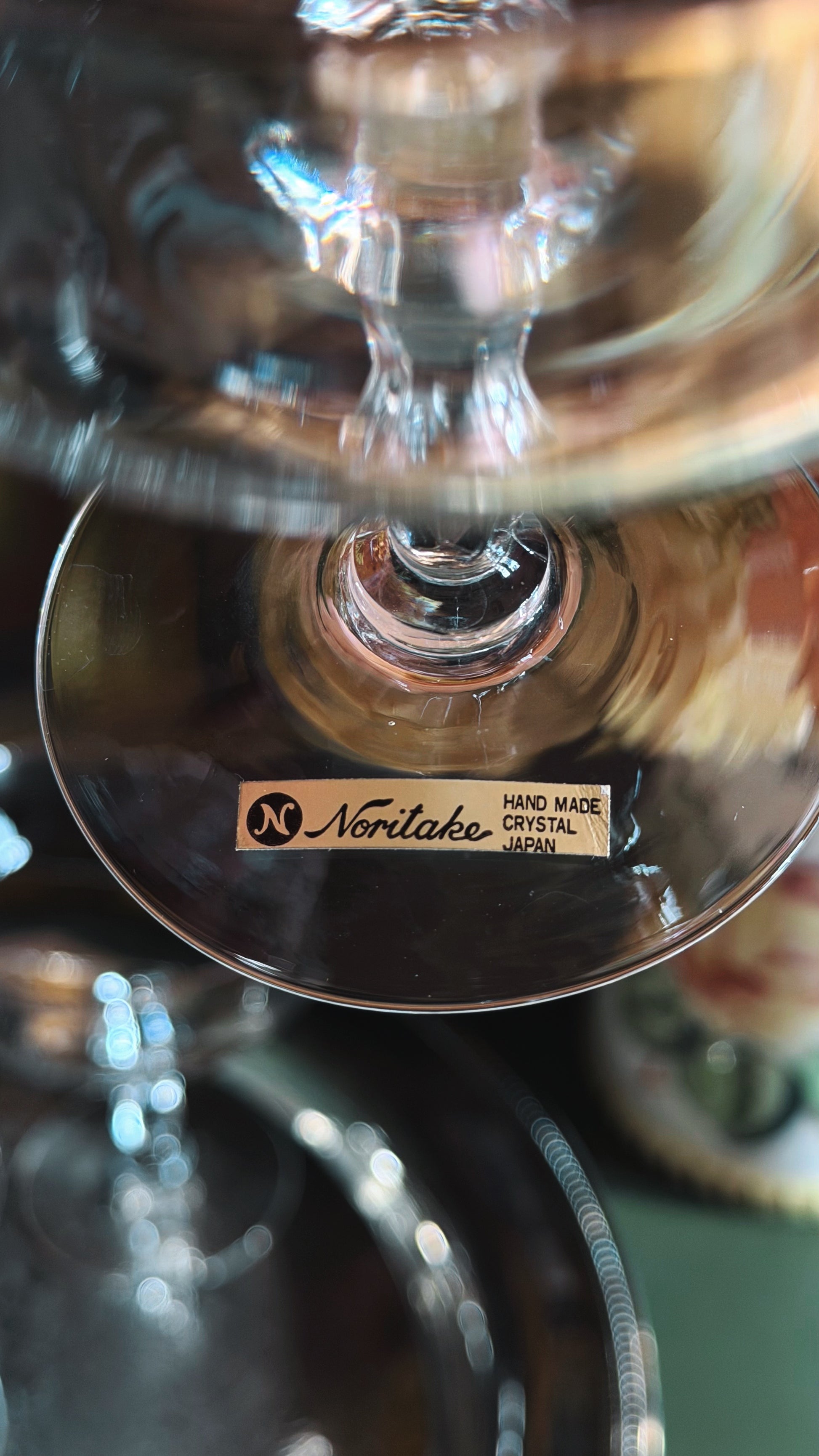 A close-up of a clear crystal champagne coupe stem with a small Noritake label on the foot, photographed against a softly blurred reflective background.