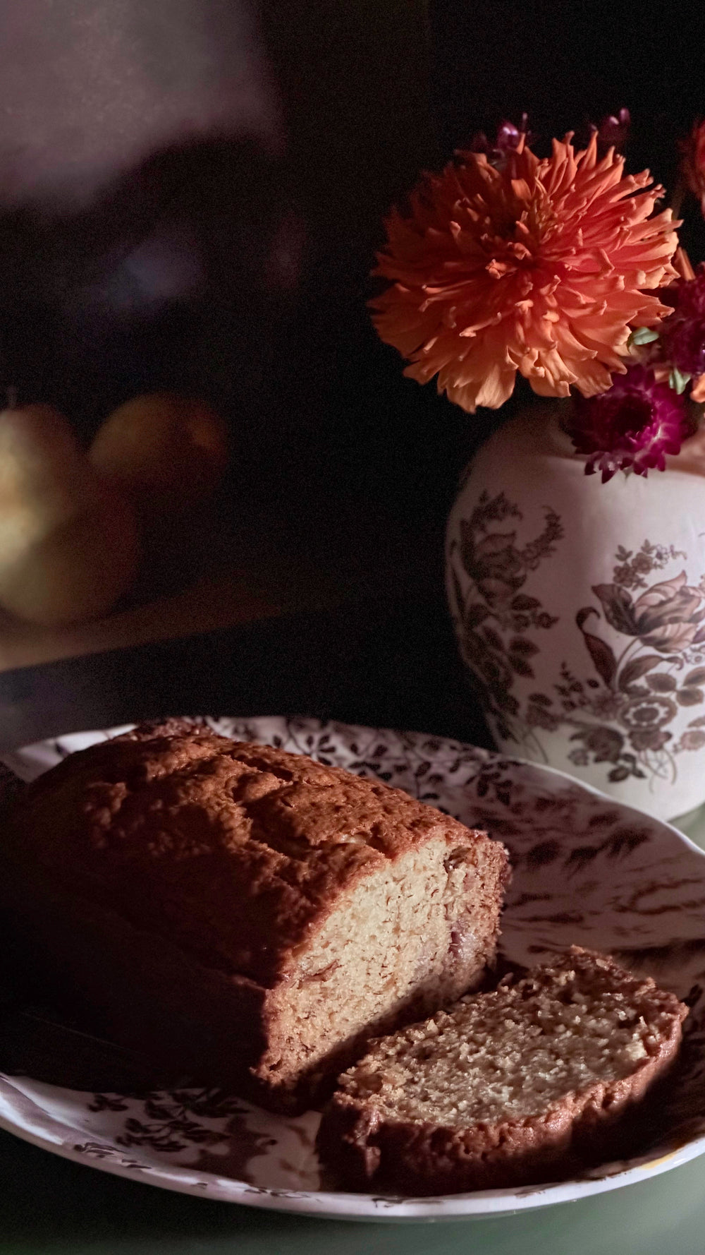 A loaf of brown bread with a slice cut, served on a patterned plate in a dimly lit setting, with a floral-patterned vase of orange flowers in the background.