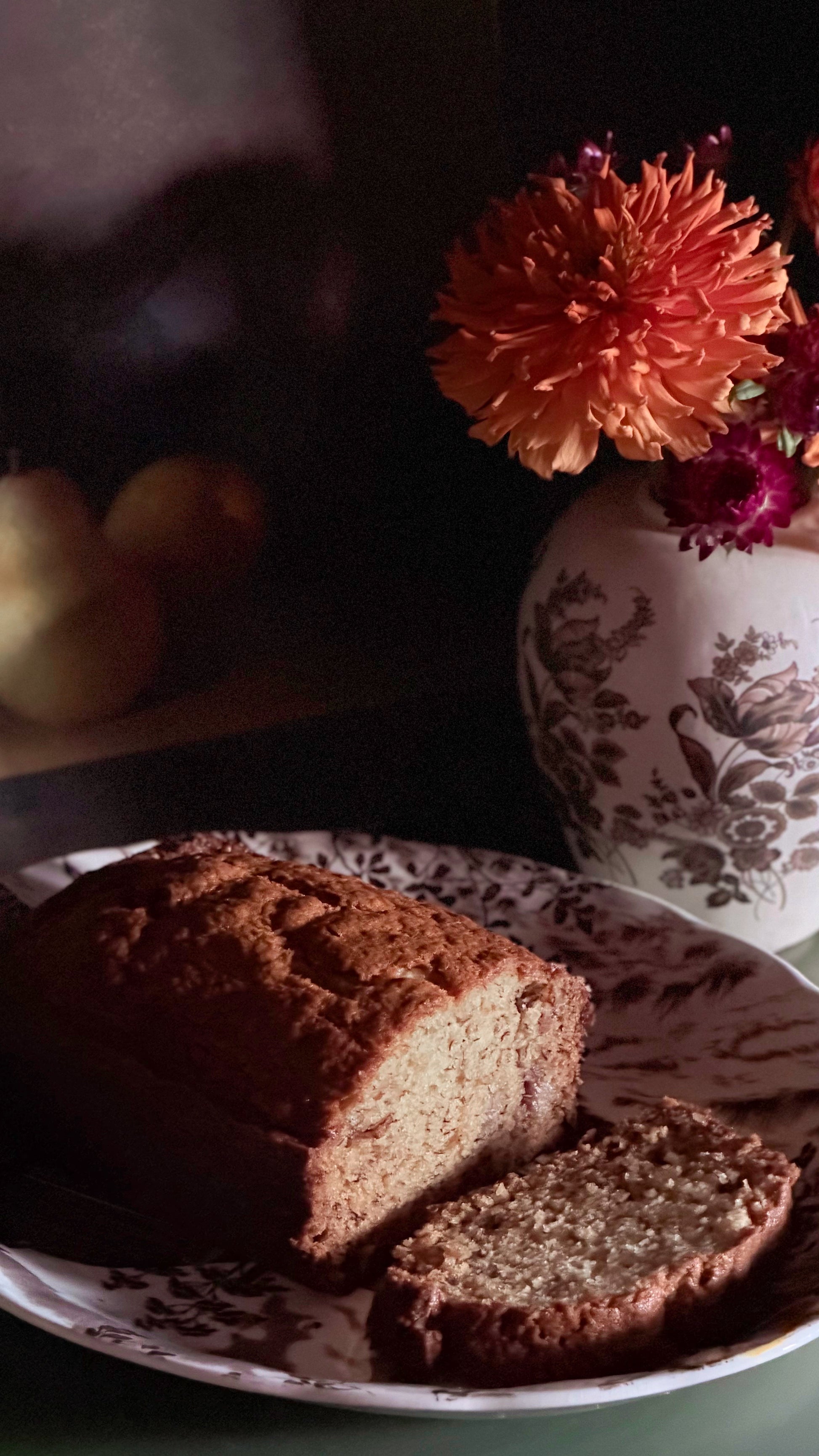 A loaf of brown bread with a slice cut, served on a patterned plate in a dimly lit setting, with a floral-patterned vase of orange flowers in the background.