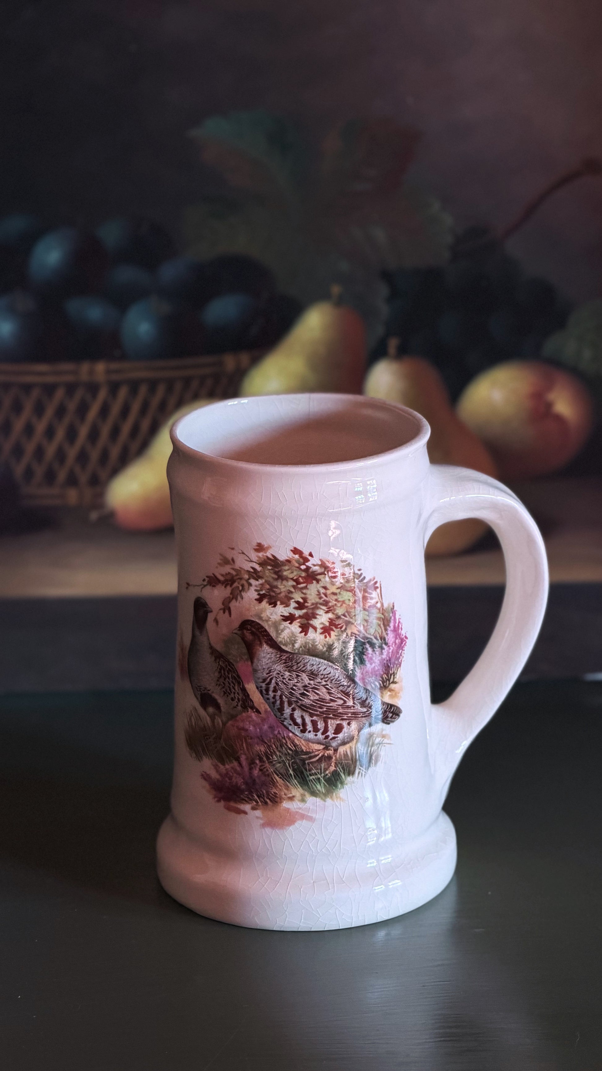 A Vintage English Pottery Game Bird Tankard | Pheasant & Partridge Mug with a handle, featuring a detailed illustration of a pheasant bird among autumn leaves and flowers on the front. The mug is set on a dark tabletop with a blurred rustic background containing grapes and pears.