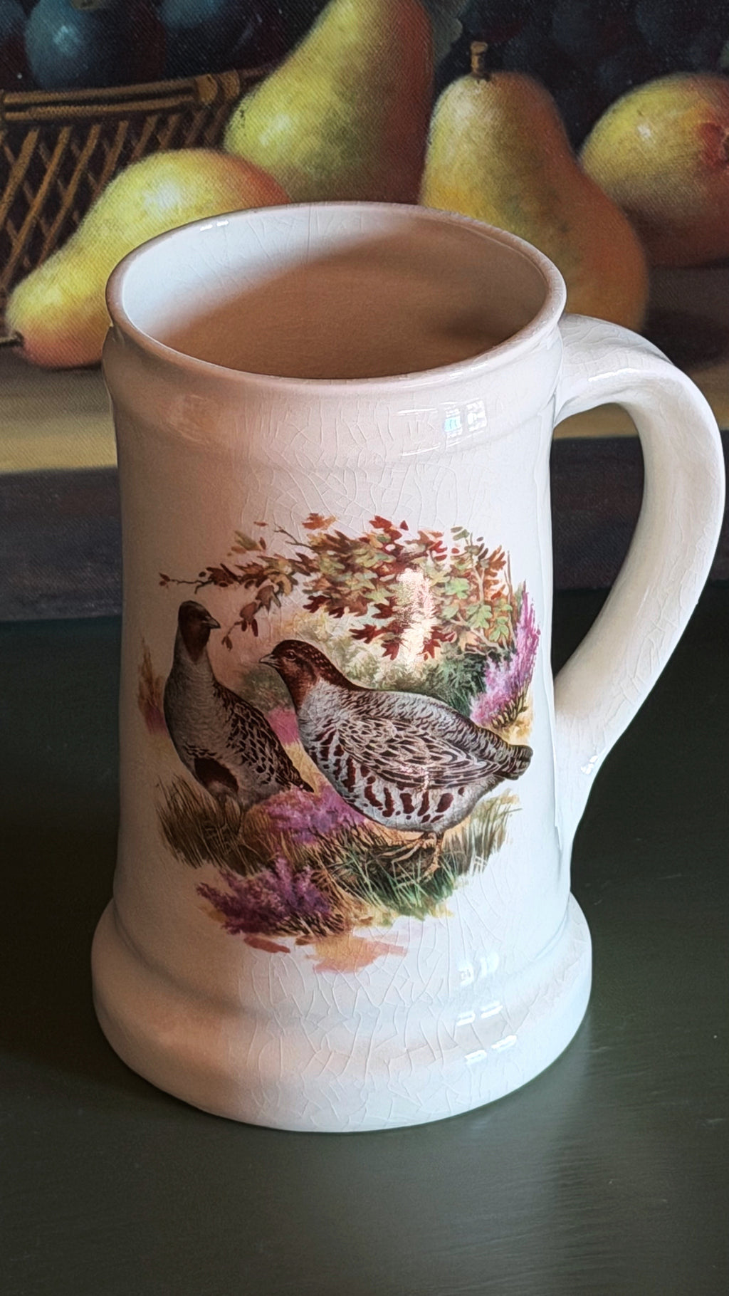 A white ceramic beer stein mug featuring a detailed illustration of a quail in a natural setting with purple flowers and autumn leaves. The mug is set on a dark tabletop with a blurred background of pears.