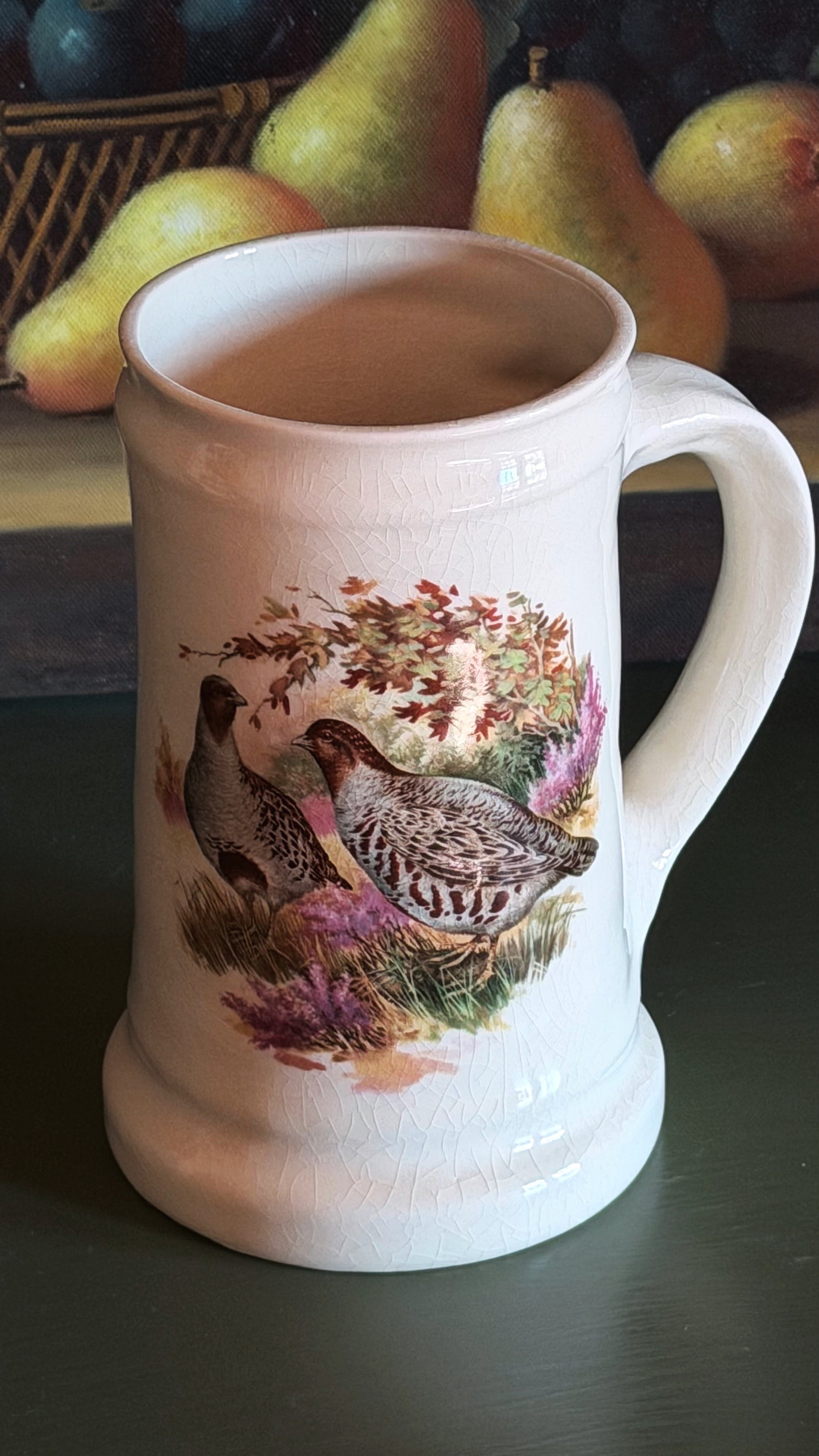 A white ceramic beer stein mug featuring a detailed illustration of a quail in a natural setting with purple flowers and autumn leaves. The mug is set on a dark tabletop with a blurred background of pears.