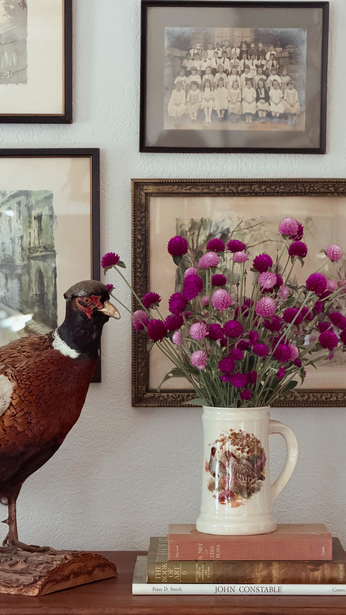 A Vintage English Pottery Game Bird Tankard filled with pink and purple flowers sits on a wooden surface next to a taxidermy pheasant, with framed vintage photographs on a wall behind it.