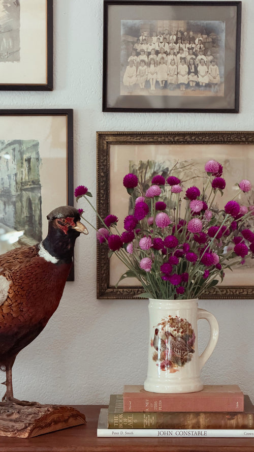 A Vintage English Pottery Game Bird Tankard filled with pink and purple flowers sits on a wooden surface next to a taxidermy pheasant, with framed vintage photographs on a wall behind it.