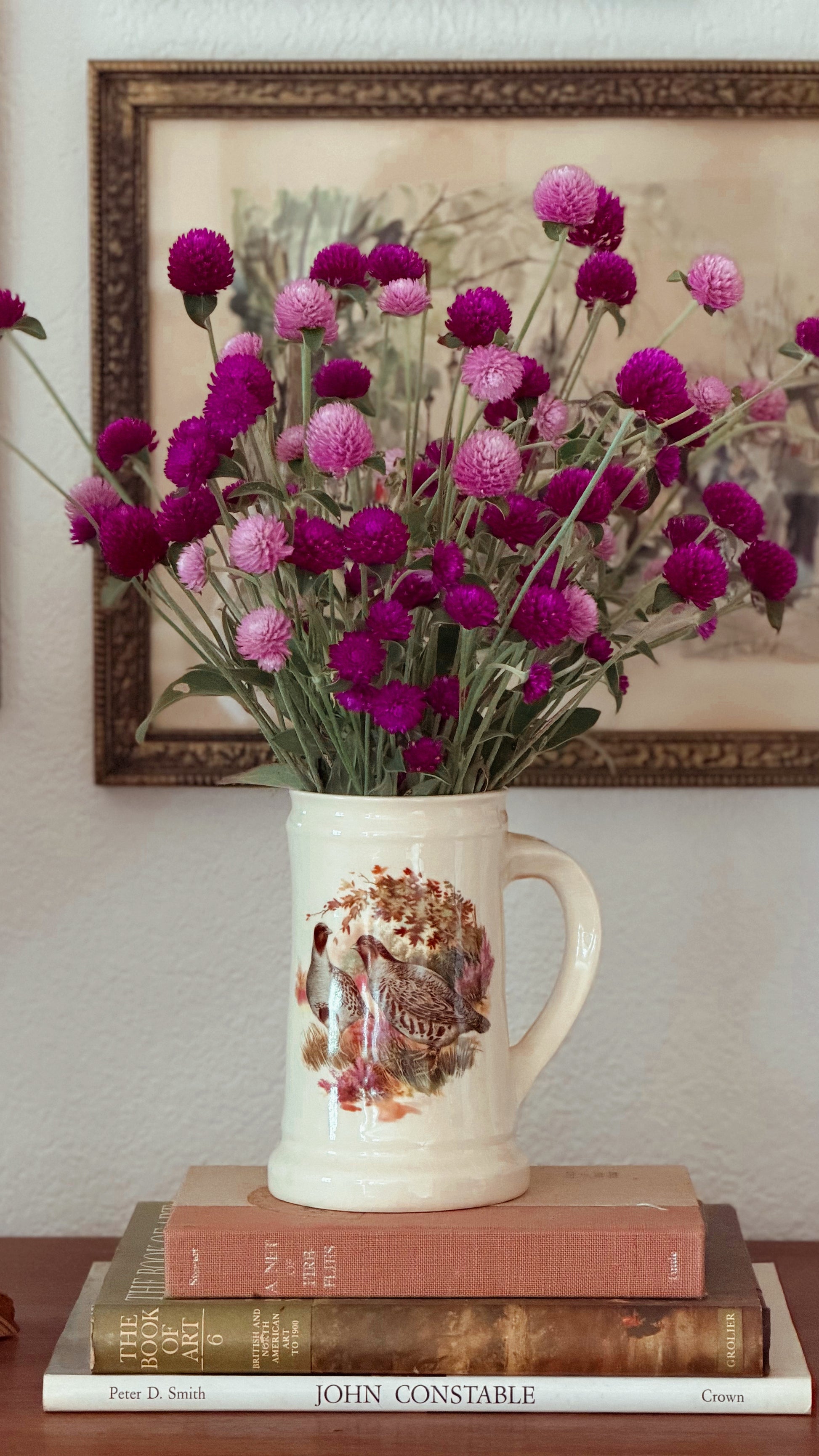 A cream-colored ceramic mug with a bird illustration sits on a stack of three books, holding a large bouquet of pink and purple globe flowers. A framed picture hangs on the wall behind.