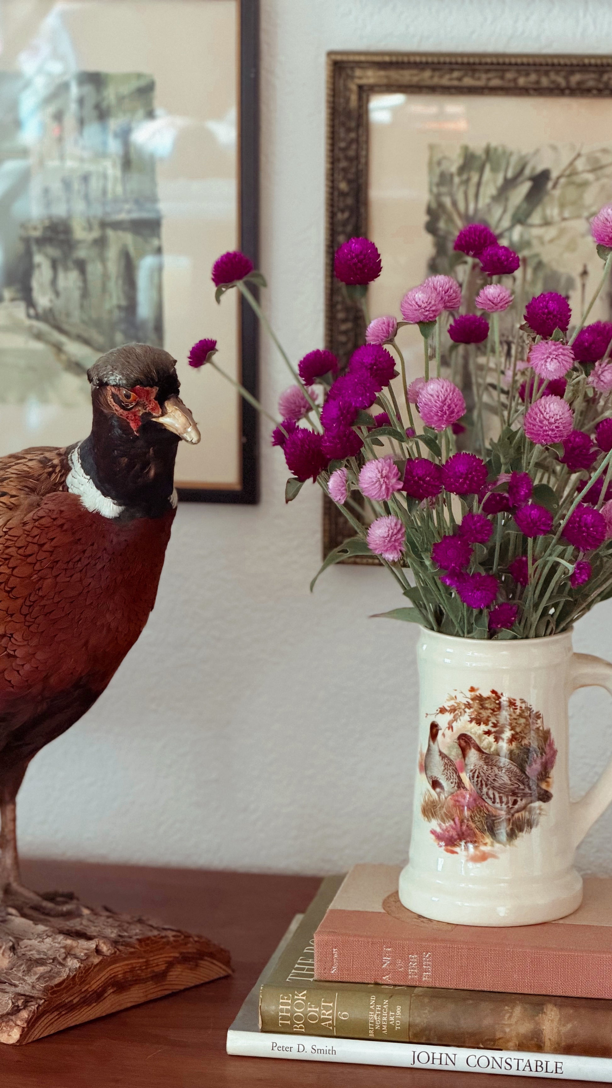A ceramic mug with a vintage-style bird illustration sits on a stack of books on a wooden table, filled with pink and purple globe flowers. A taxidermy pheasant is standing on the left, and framed wall art hangs on the wall behind.
