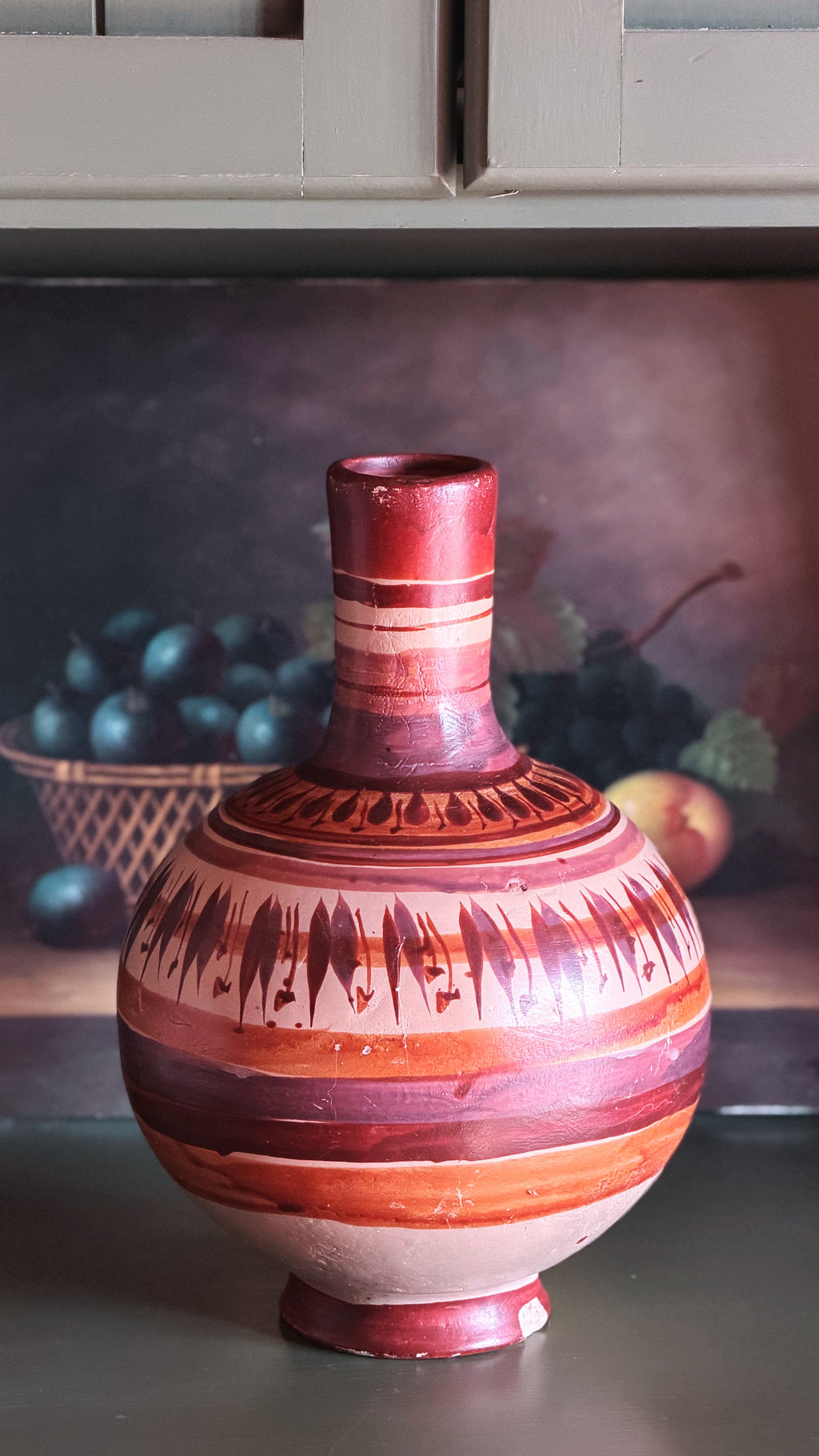 A decorative ceramic vase with a bulbous body, narrow neck, and footed base, decorated with horizontal bands of terracotta red, rust orange, and cream, along with dark brown geometric patterns near the shoulder. It is set against a dark, moody background with a blurred still life of grapes and fruit.