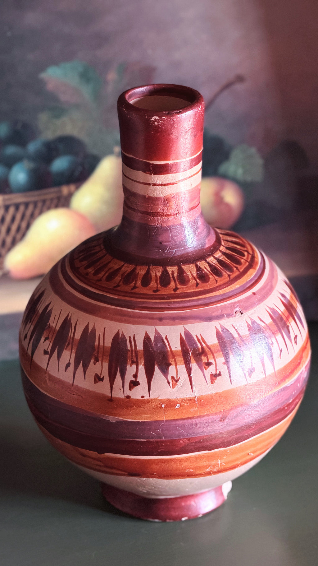 A decorative ceramic vase with a rounded body, narrow neck, and footed base, decorated with horizontal bands and geometric patterns in reddish-brown, terracotta, and cream, set against a softly blurred background.