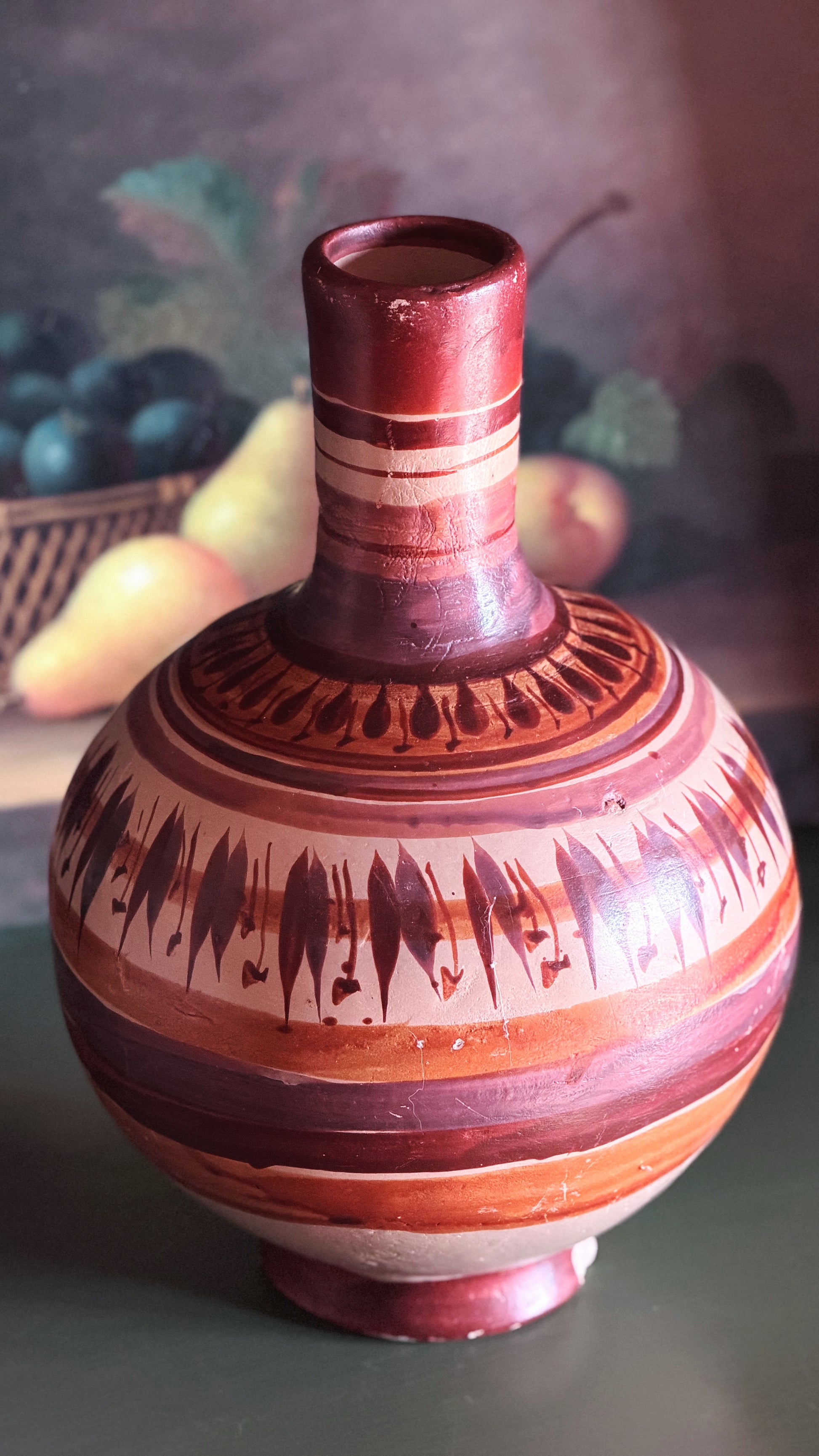 A decorative ceramic vase with a rounded body, narrow neck, and footed base, decorated with horizontal bands and geometric patterns in reddish-brown, terracotta, and cream, set against a softly blurred background.