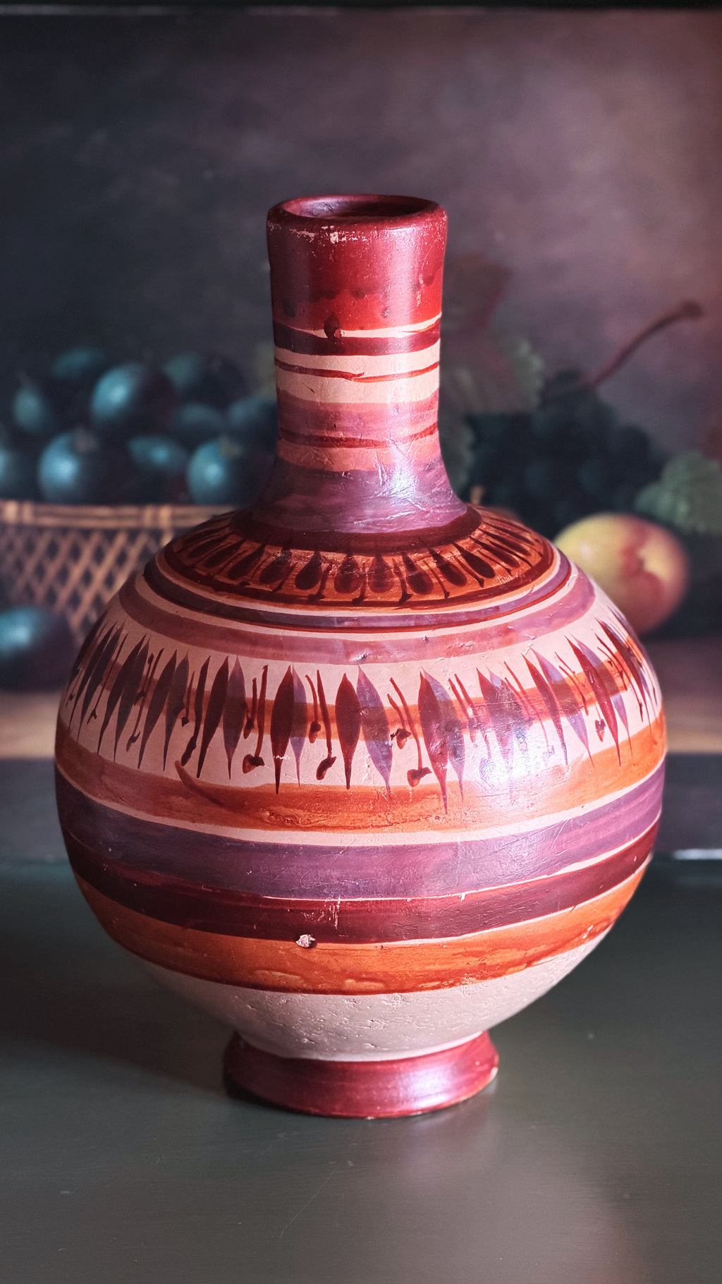 A hand-painted ceramic vase with a bulbous body and narrow neck, decorated with horizontal bands of terracotta, reddish-brown, and cream, along with black geometric and fish-like patterns, set against a dark, softly blurred background.