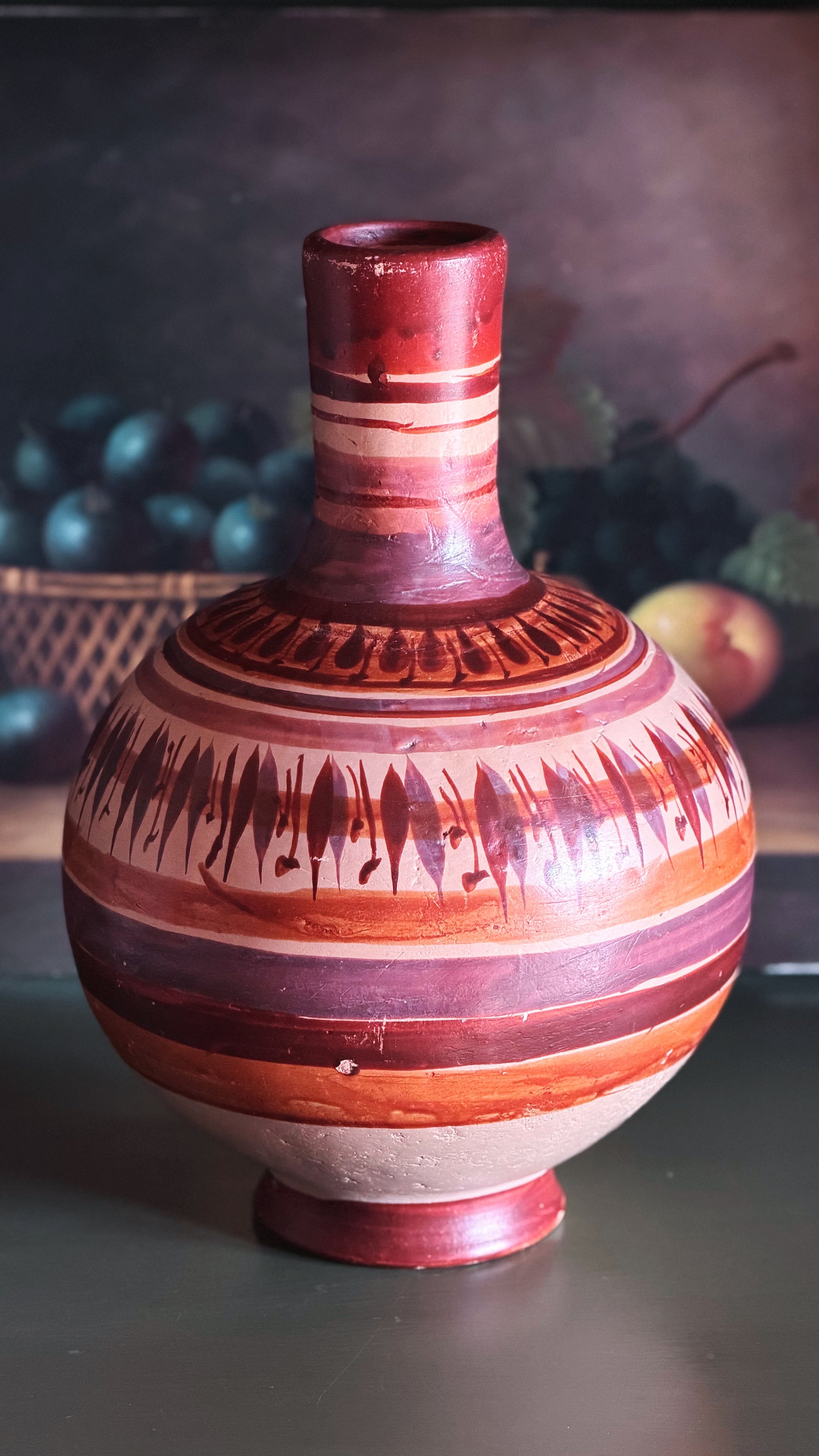 A hand-painted ceramic vase with a bulbous body and narrow neck, decorated with horizontal bands of terracotta, reddish-brown, and cream, along with black geometric and fish-like patterns, set against a dark, softly blurred background.