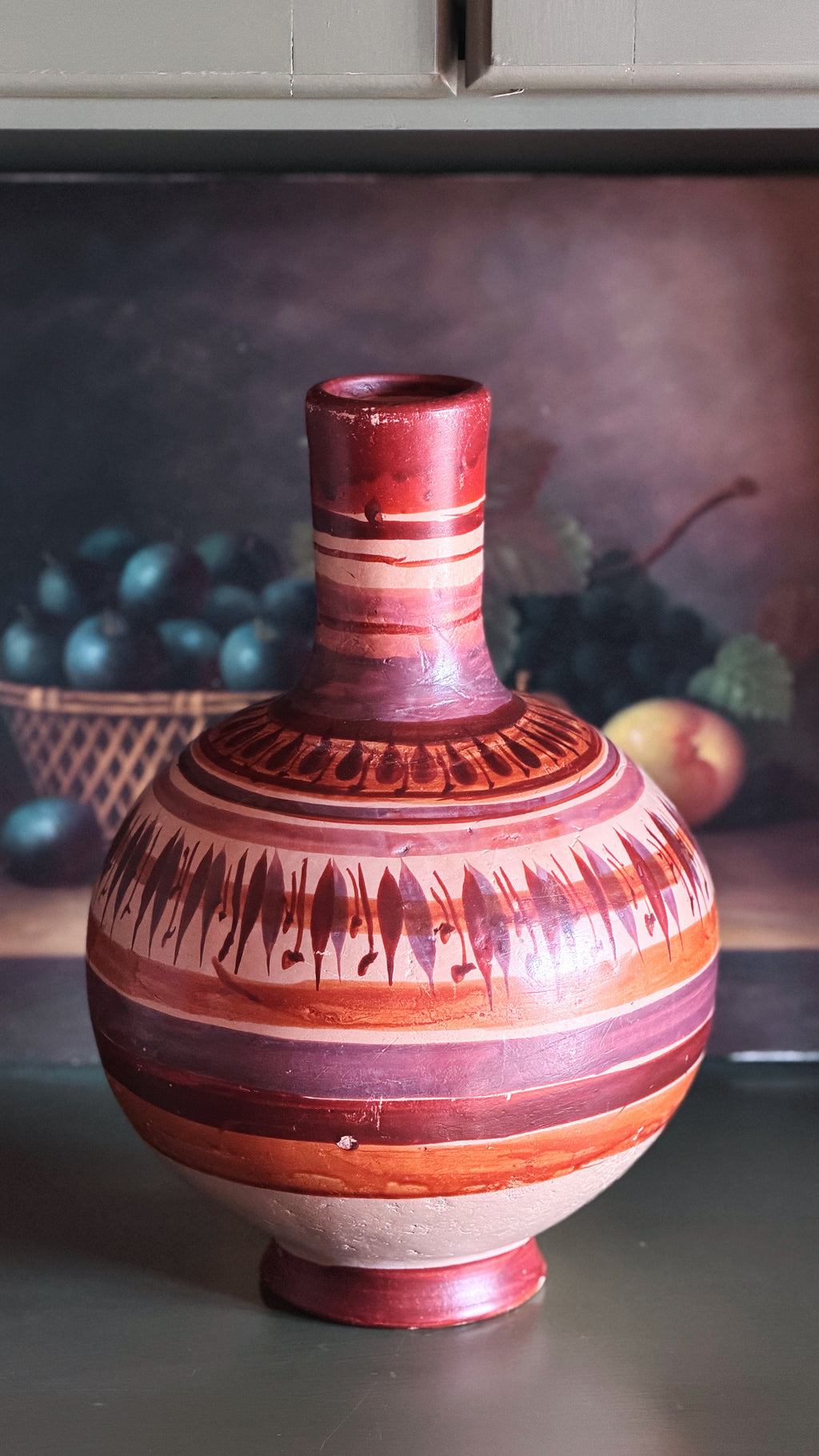 A decorative ceramic vase with a bulbous body, narrow neck, and footed base, featuring horizontal bands of terracotta red, rust orange, and cream with painted geometric and leaf-like motifs.