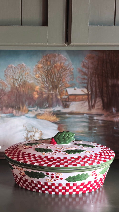 A round lidded ceramic dish or tureen with a red and white checkered pattern, green holly leaves, and a small green leaf lid handle, set on a countertop in front of a painted winter landscape.