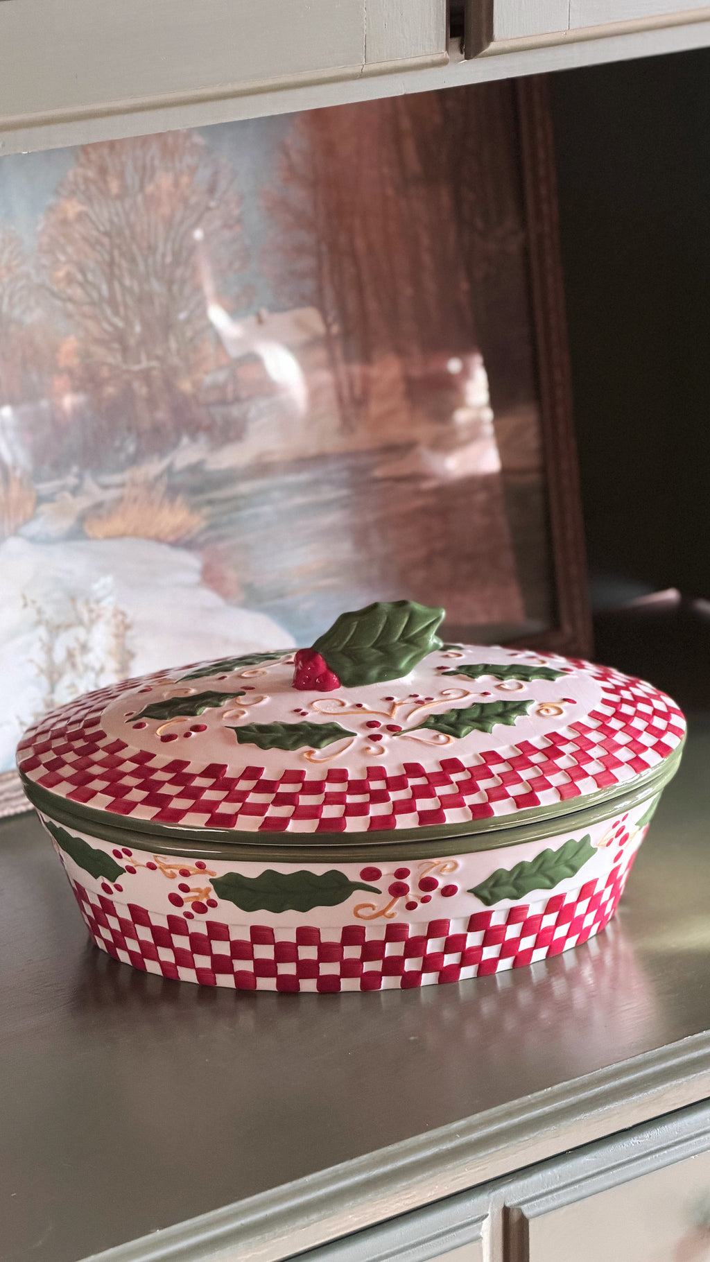 A Vintage Temp-tations by Tara Winter Holly 2 Quart Covered Casserole Dish with a red and white checkered pattern, green holly leaf designs, and a small green leaf topper with a red berry, sitting on a metallic countertop.