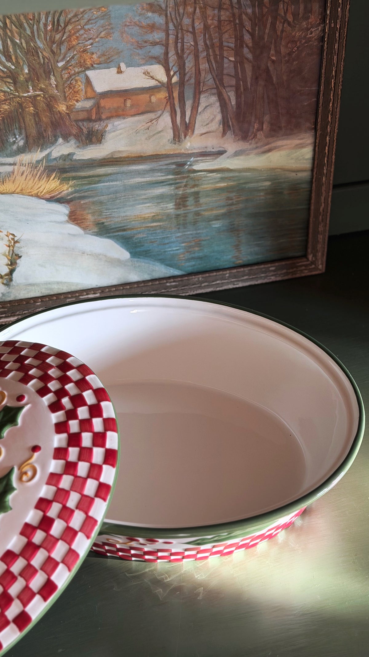 A round white ceramic baking dish with a red and white checkered patterned rim and matching lid, shown on a dark tabletop with a framed winter landscape painting in the background.