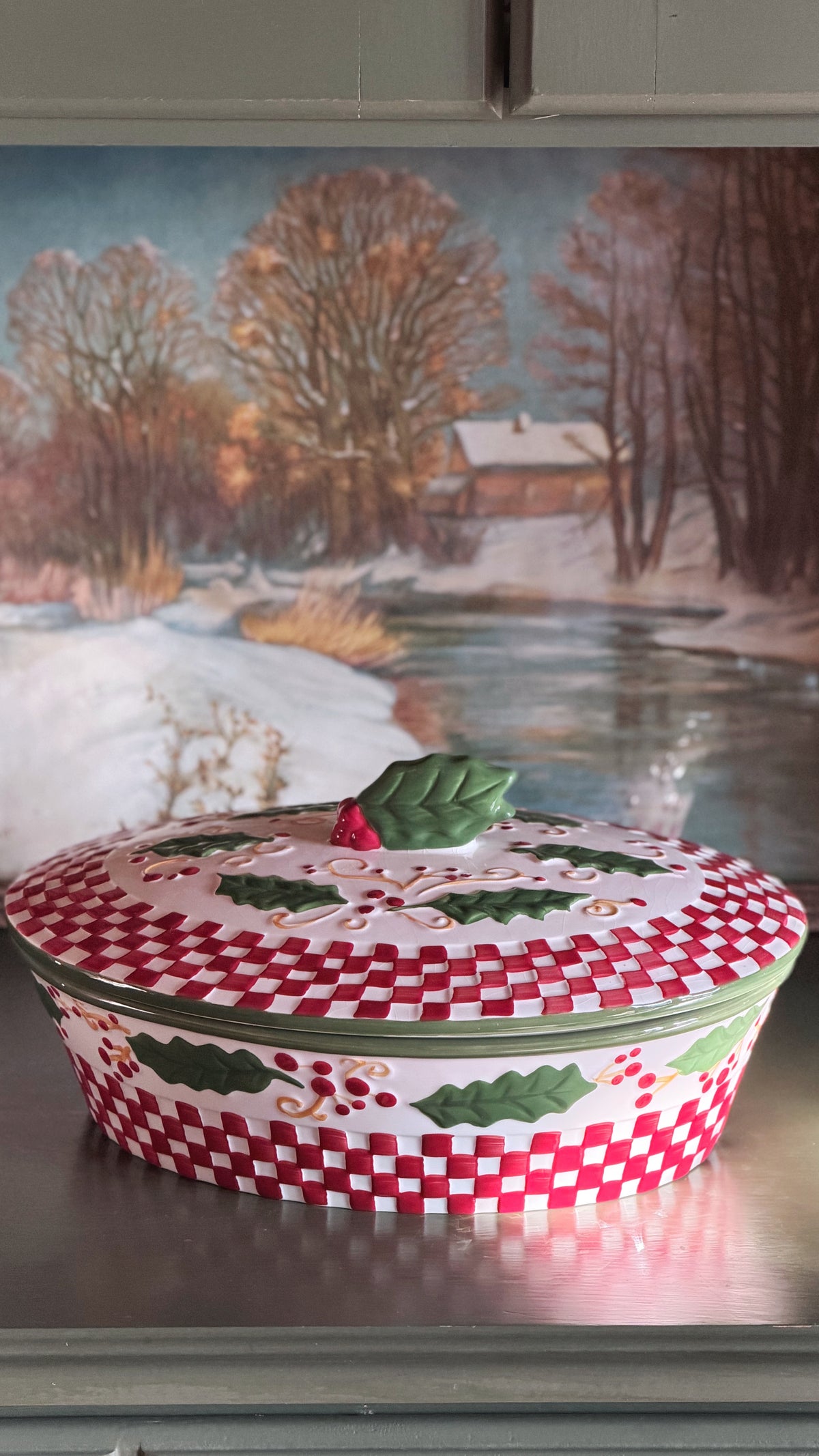 A Vintage Temp-tations by Tara Winter Holly 2 Quart Covered Casserole Dish with a red and white checkered pattern, green holly leaves, and small red berries, set against a painted winter landscape background.