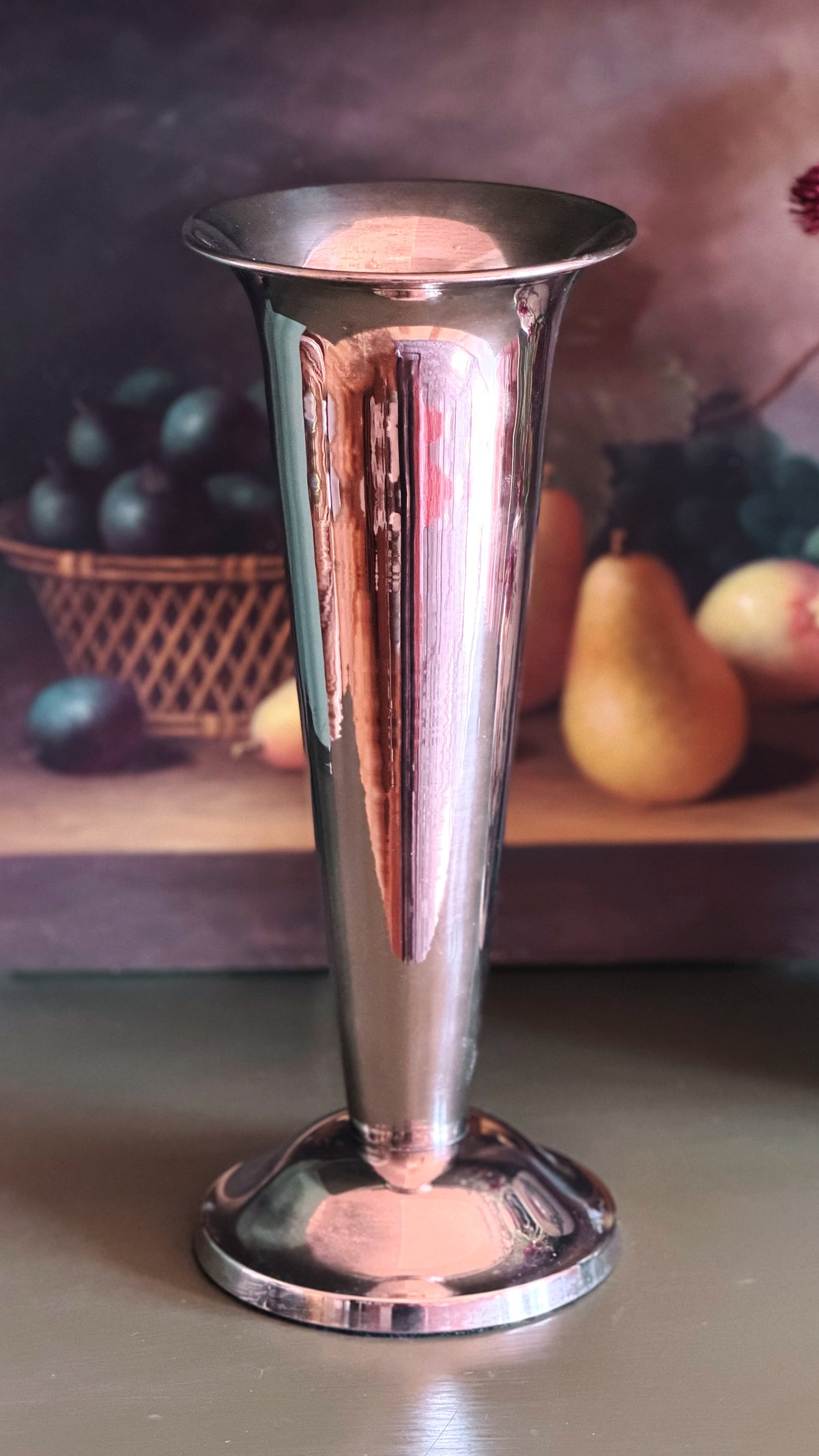 A polished silver metal flared vase or goblet-shaped vessel with a reflective finish, standing on a round base against a softly blurred rustic background with fruit.