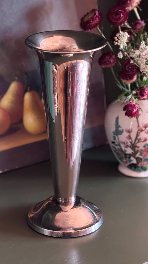 A polished silver-toned metal flared vase or goblet with a circular base, shown on a tabletop with a blurred floral arrangement and fruit in the background.